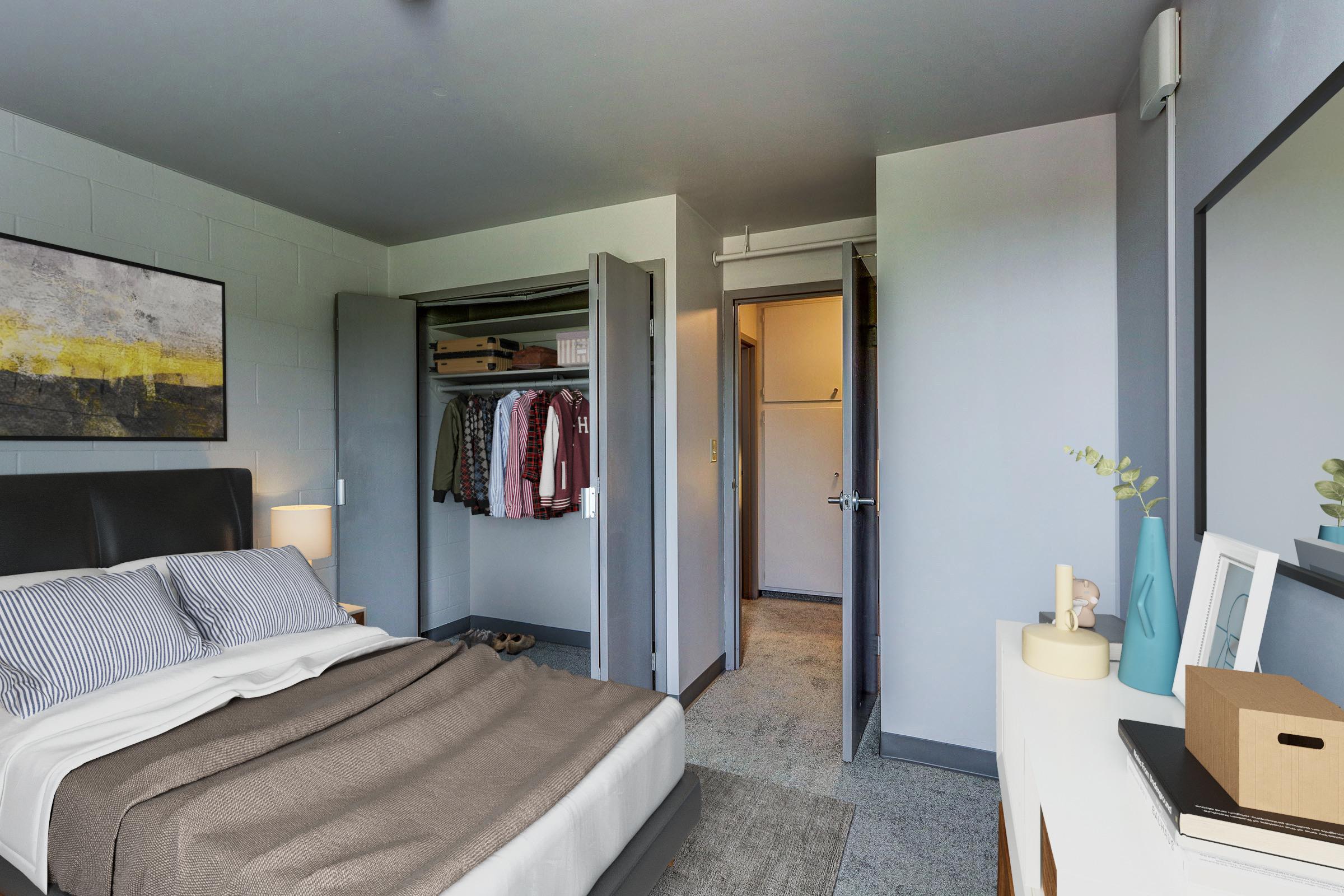 A cozy bedroom featuring a plush bed with gray bedding, a large painting on the wall, and a neatly organized closet with clothes visible. A soft rug covers the floor, and a light-colored dresser holds decorative items. A doorway leads to a small hallway or bathroom area.
