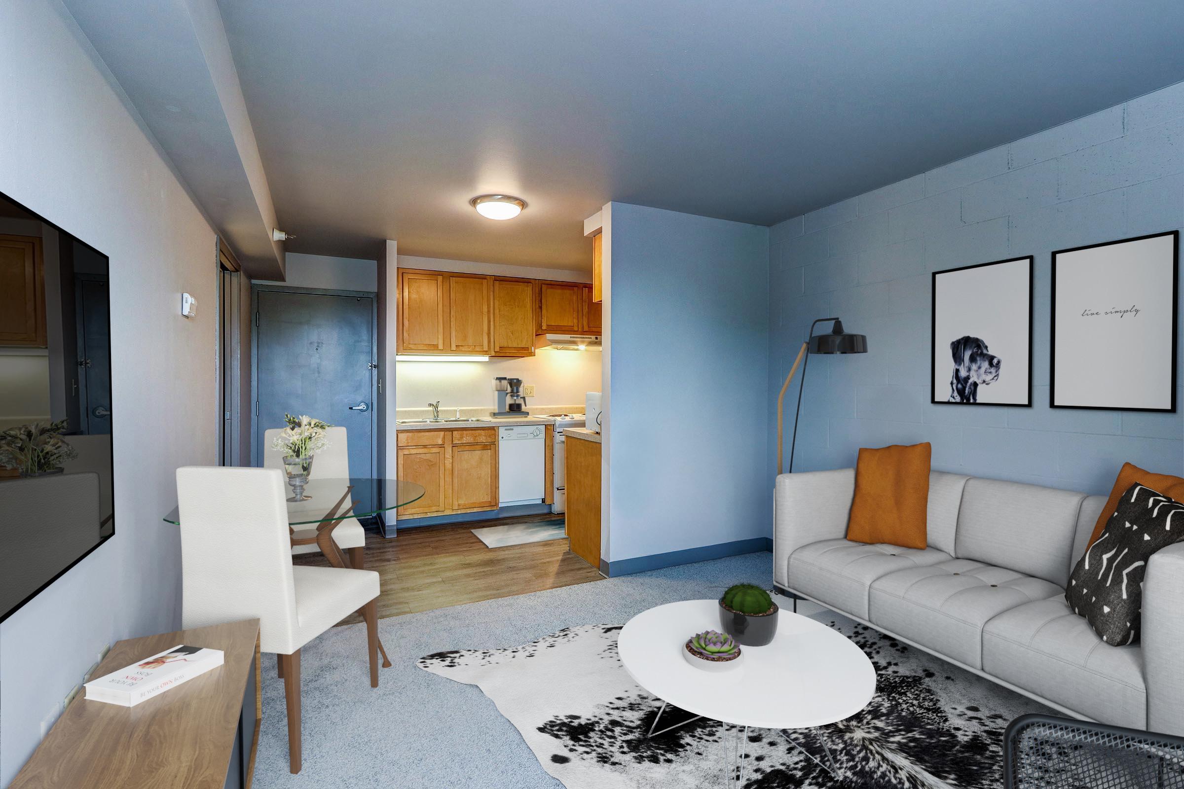 A modern living room featuring a light gray sofa with orange and patterned cushions, a white coffee table on a black and white rug, and a wall-mounted TV. In the background, an open kitchen with wooden cabinets and appliances is visible, along with a door leading to another room. Decor includes framed artwork and a plant.