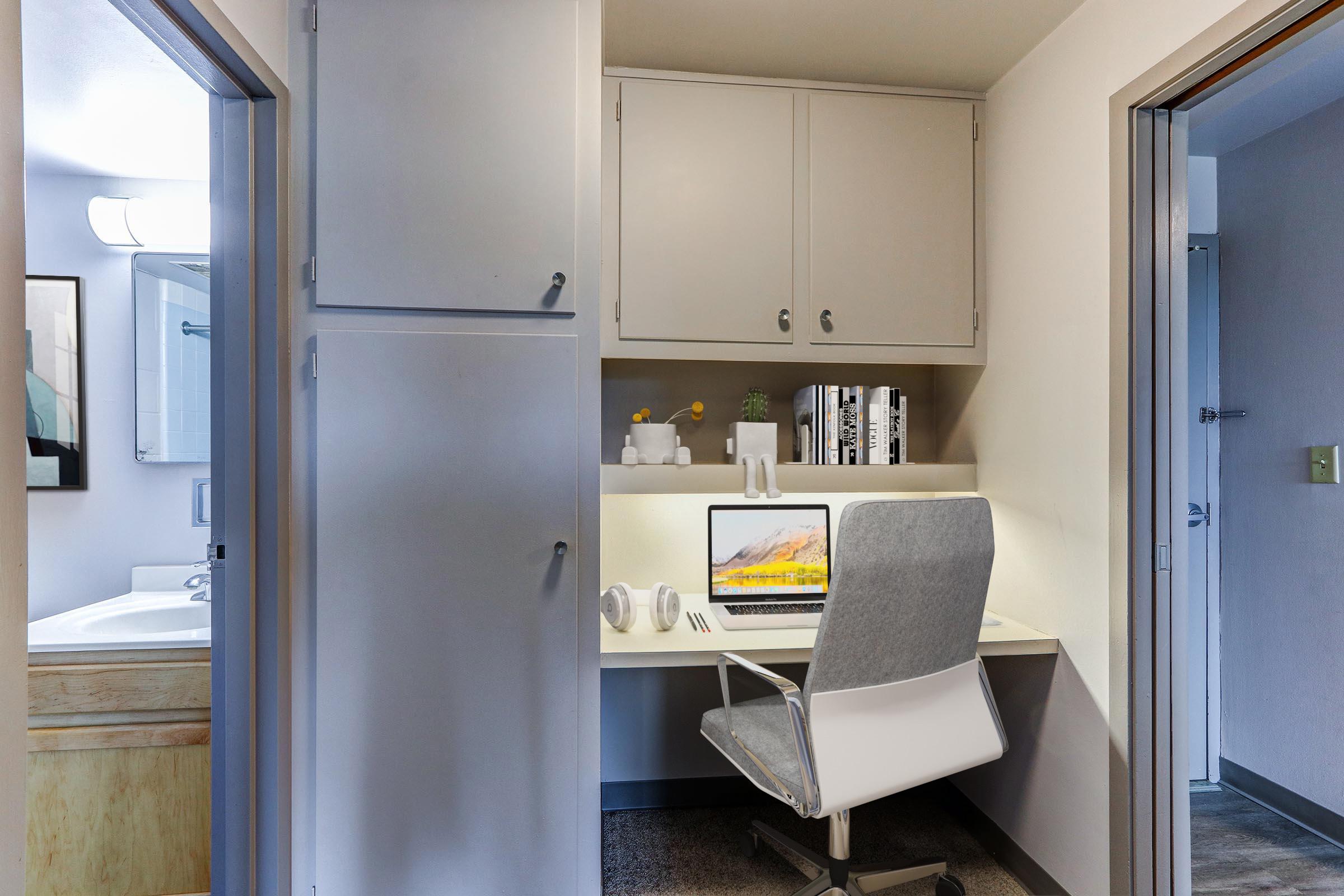 A compact workspace featuring a computer on a desk with a gray chair, surrounded by cabinets. The area is well-lit, with a small vase of flowers and books on the shelf. In the background, a bathroom with a mirror and sink is visible, along with a door leading to another room.