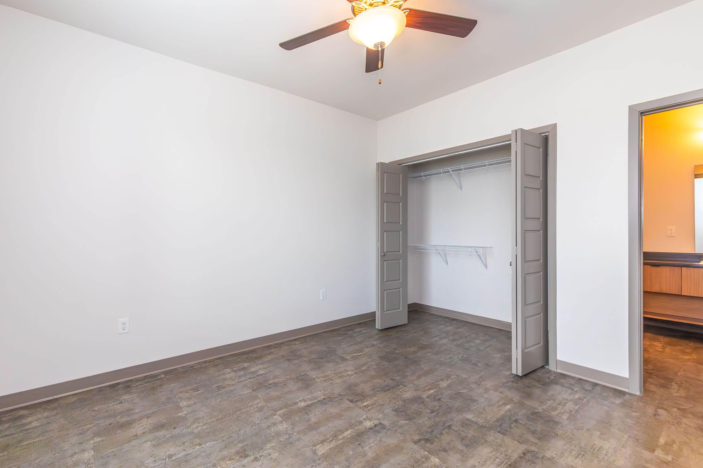 An empty room with light-colored walls and a ceiling fan. There is a closet with double doors and shelves inside, located on the left side. The flooring is a wood-like material, and a doorway on the right leads to another space, partially visible. Natural light enters from the adjacent room.