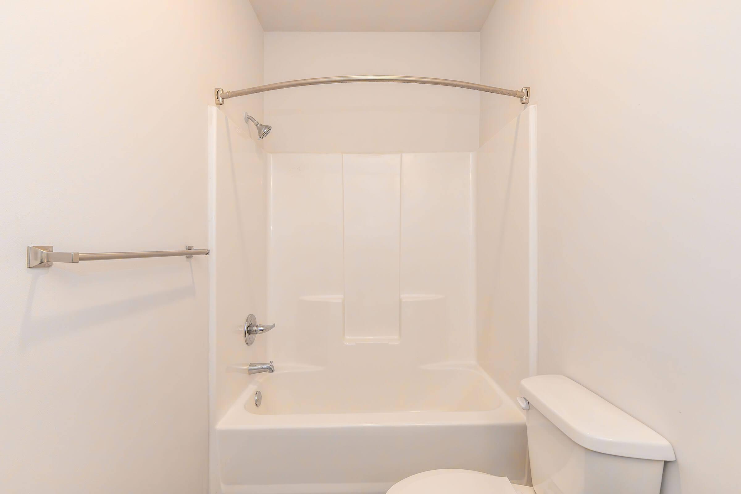 A clean and minimalist bathroom featuring a white bathtub with a showerhead, a chrome grab bar, and a simple white toilet. The walls are plain white, creating a bright and airy atmosphere in the space.