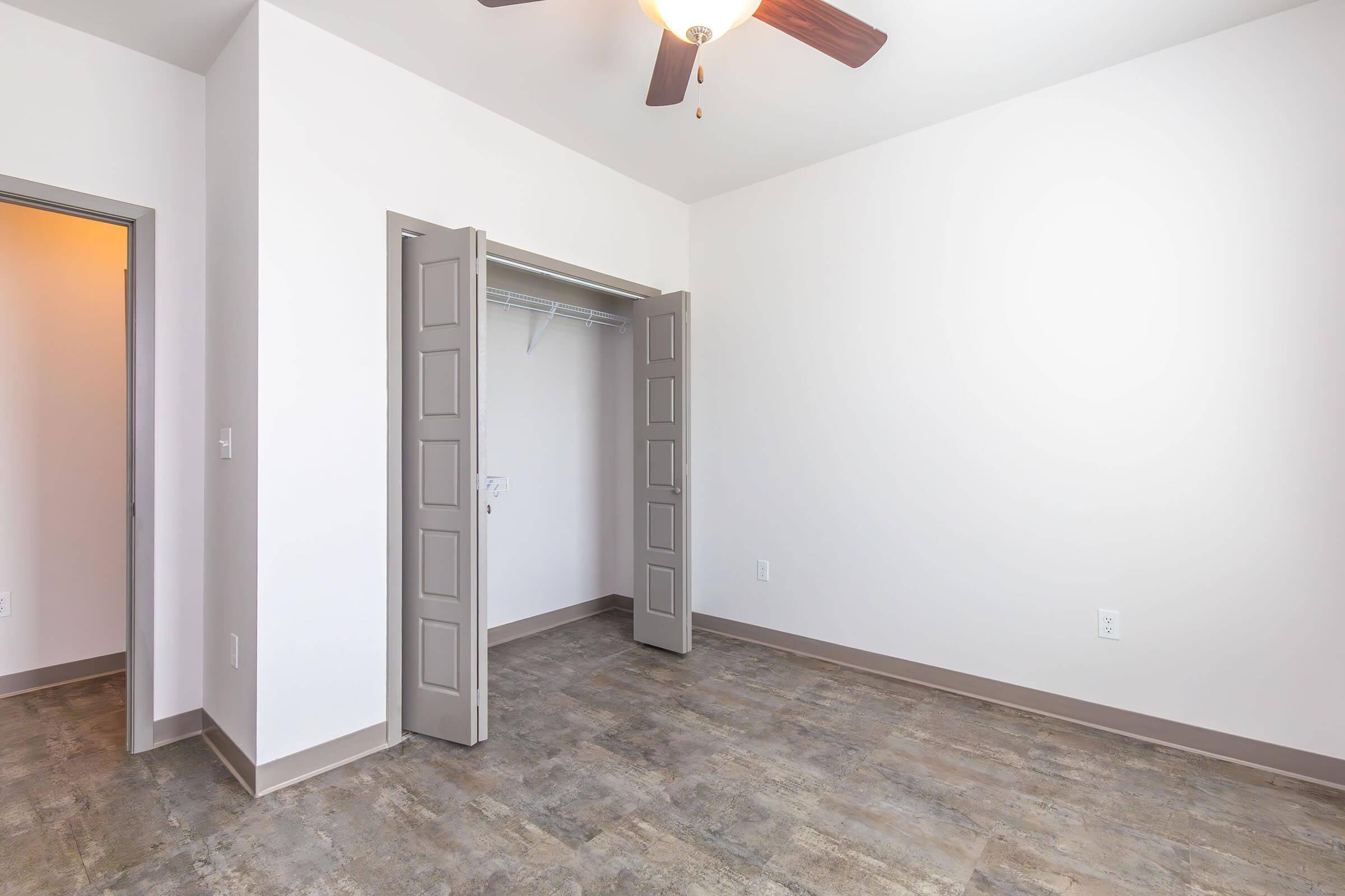 A spacious, empty room featuring light-colored walls and a ceiling fan. There are two open doors leading to a closet on the left side, and the floor is covered with a light-colored tile. Natural light fills the room, creating a bright and airy atmosphere.