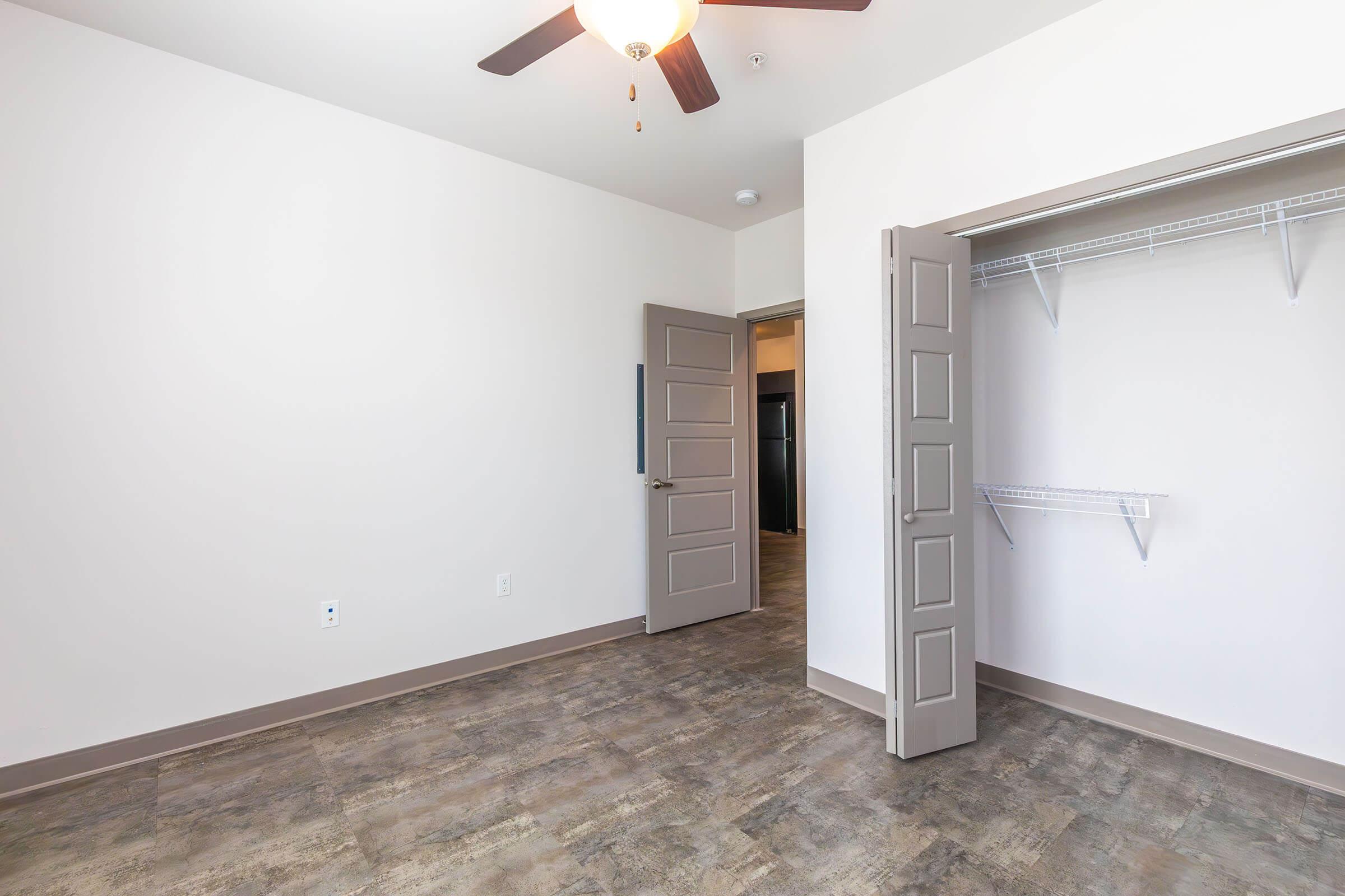 A clean, empty room with a ceiling fan, light-colored walls, and a closet with shelves. The floor features a modern, textured design. A doorway leads to another room, and the overall space appears bright and well-maintained.
