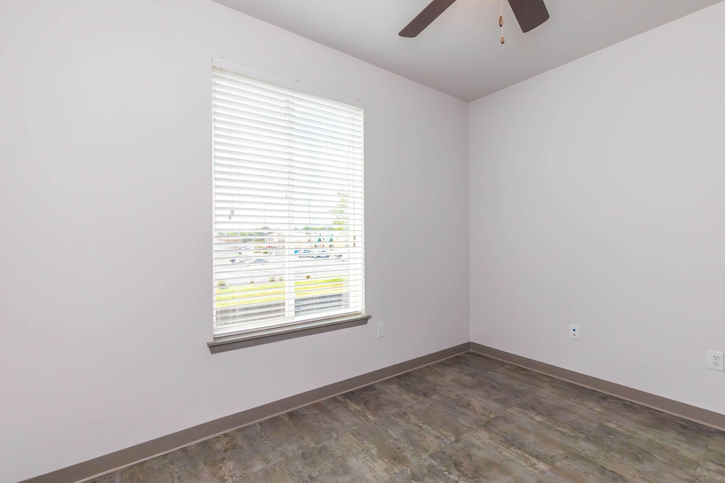 Empty room with light gray walls and a large window dressed with white blinds. The floor is a wood-like laminate, and there is a ceiling fan with wooden blades. The space appears bright and offers a view outside through the window, which is positioned in the corner of the room.