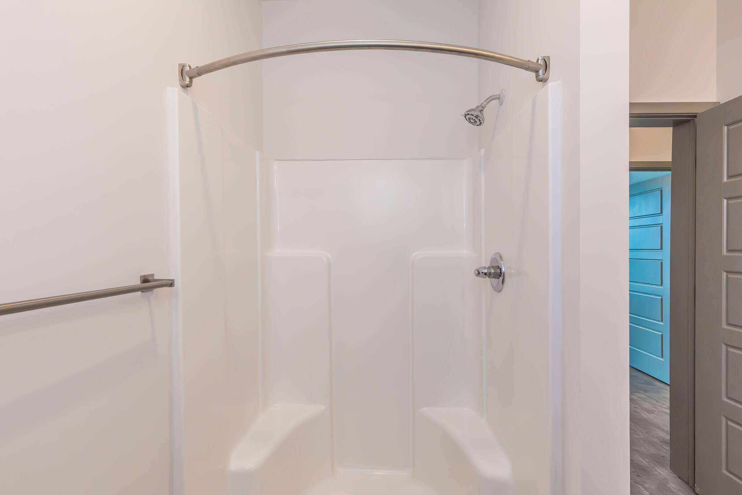 A clean, empty shower stall with a curved shower rod and a handheld showerhead. The walls are white and smooth, with two built-in benches for seating. A doorframe is visible in the background, leading to another room with a light blue door. The floor appears to be tiled in a light color.