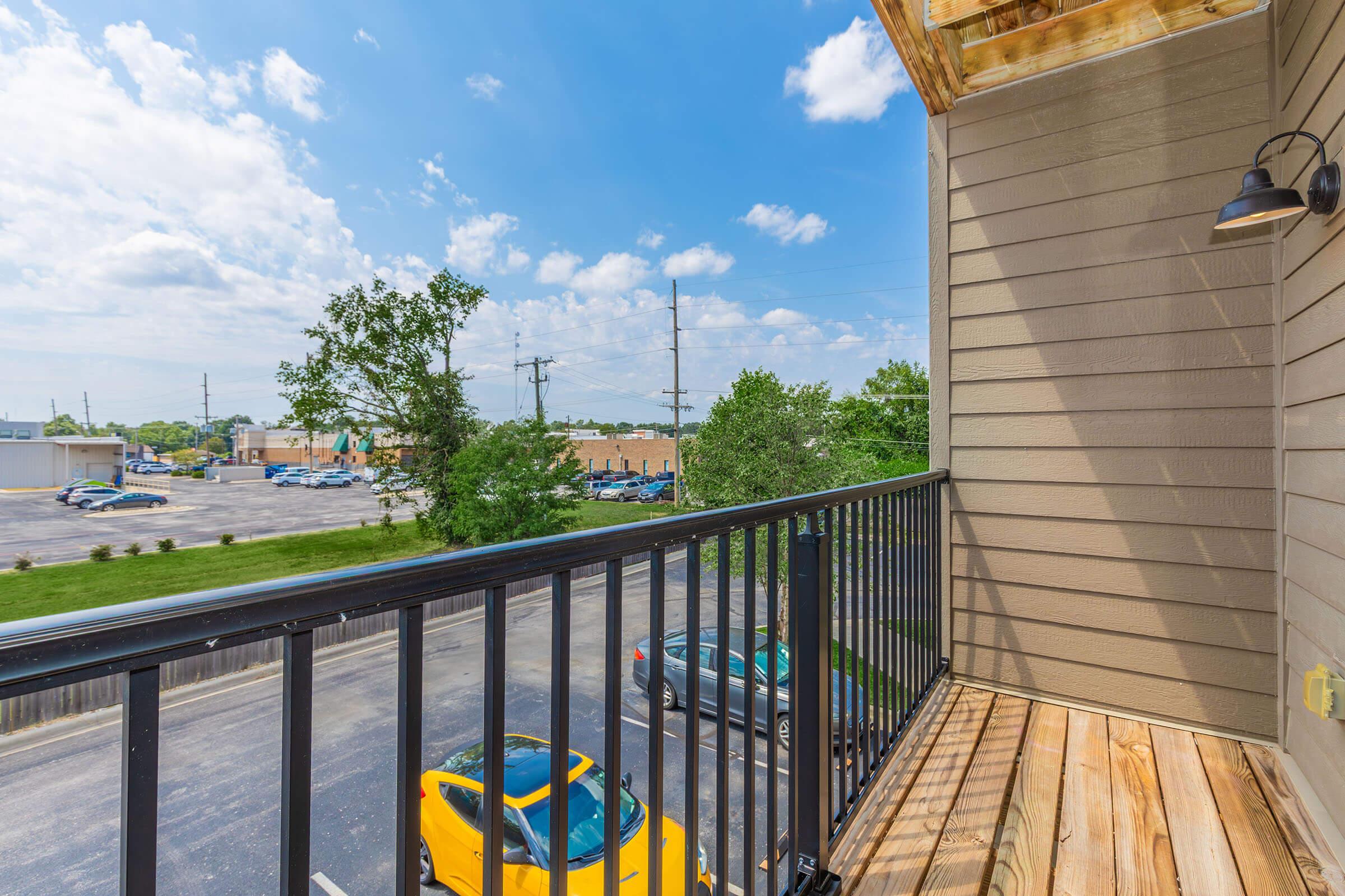 A view from a balcony featuring a wooden deck and black railing, overlooking a parking lot with a yellow car parked below. In the distance, there are commercial buildings and a few trees under a bright blue sky with scattered clouds.