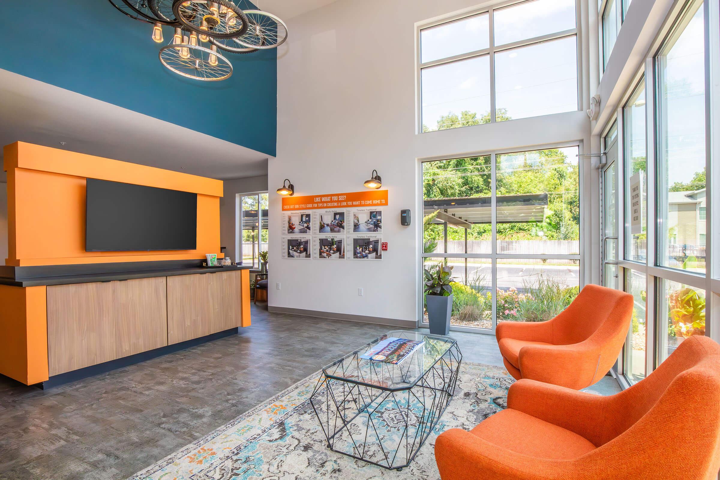 A modern reception area featuring an orange and gray color scheme. There is a sleek front desk with a large screen, a glass coffee table, and two bright orange chairs. Natural light streams through large windows, and a wall displays framed photos. Stylish decor includes pendant lighting and a decorative rug.