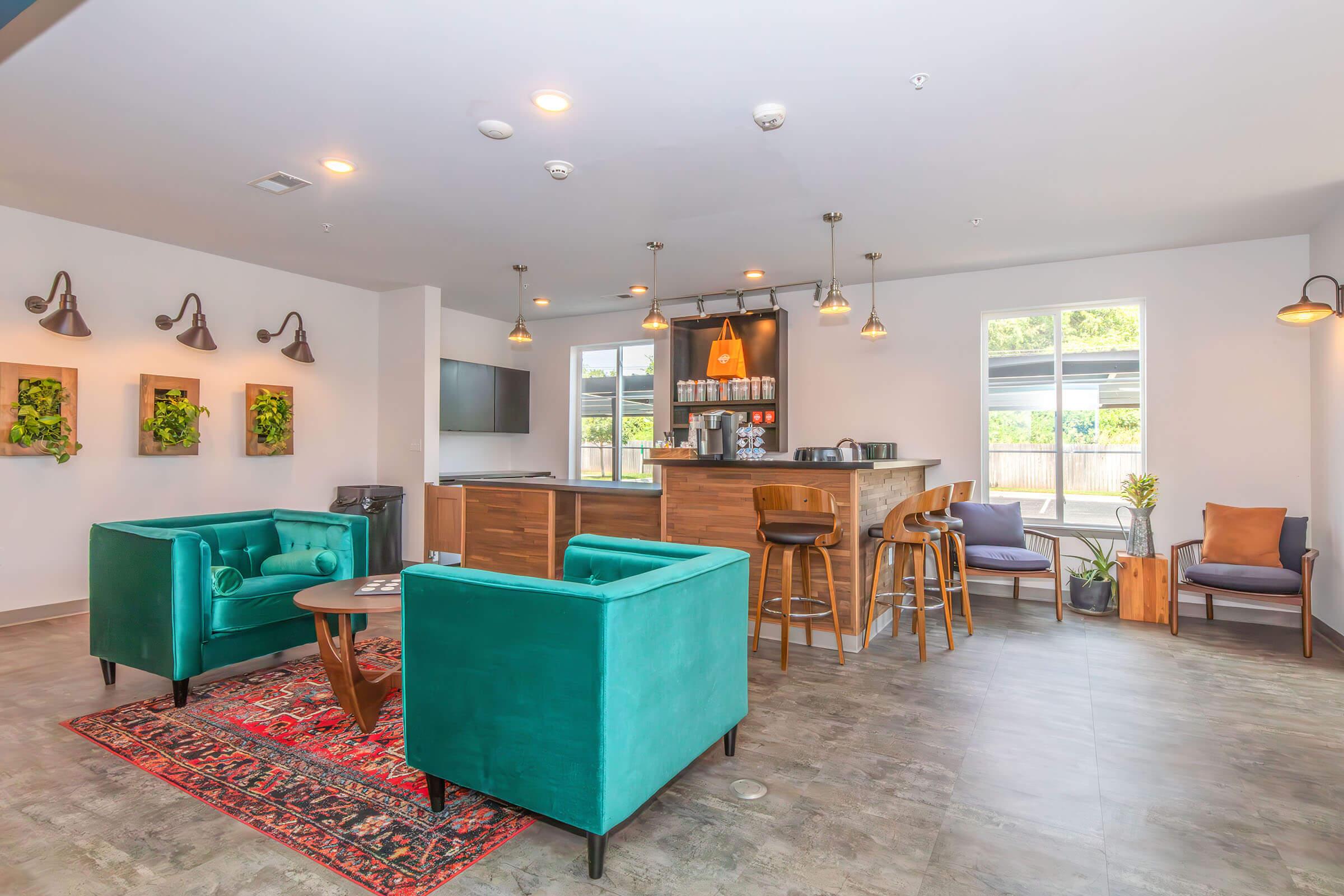 A modern lounge area featuring two teal armchairs, a wooden table, and a rustic rug. In the background, there is a stylish bar with seating, various plants on the walls, a large TV, and large windows allowing natural light to fill the space. The decor is contemporary and inviting.