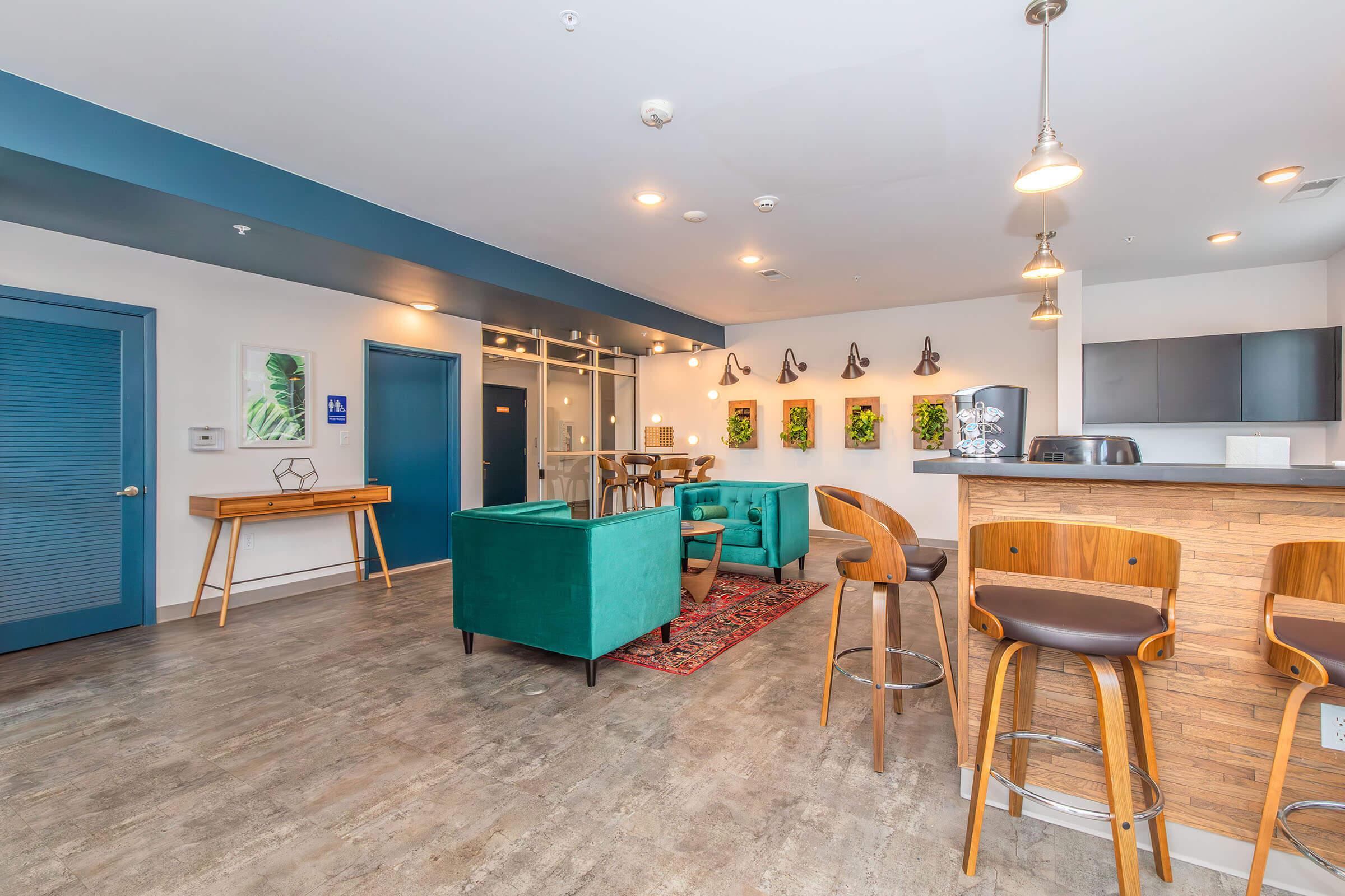 A modern lounge area with teal armchairs, wooden barstools, and a light wooden bar. The walls feature blue accents, framed green plant artwork, and decorative lighting. A glass partition separates the lounge from another space, creating an inviting and stylish atmosphere for relaxation or socializing.