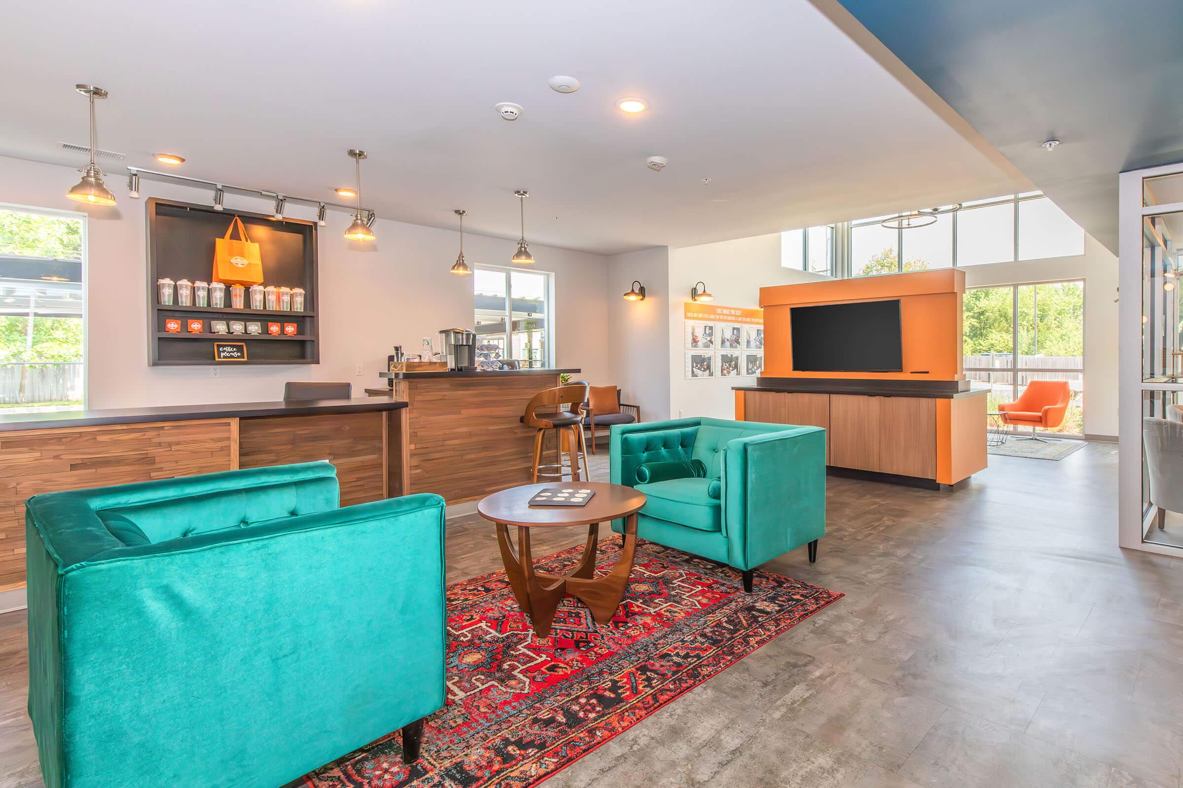 Modern lounge area with two teal armchairs, a wooden coffee table on a patterned rug, and a reception desk featuring coffee and snacks. A flat screen TV is mounted on a wall opposite large windows. The space is bright and inviting, with contemporary decor and a cozy atmosphere.