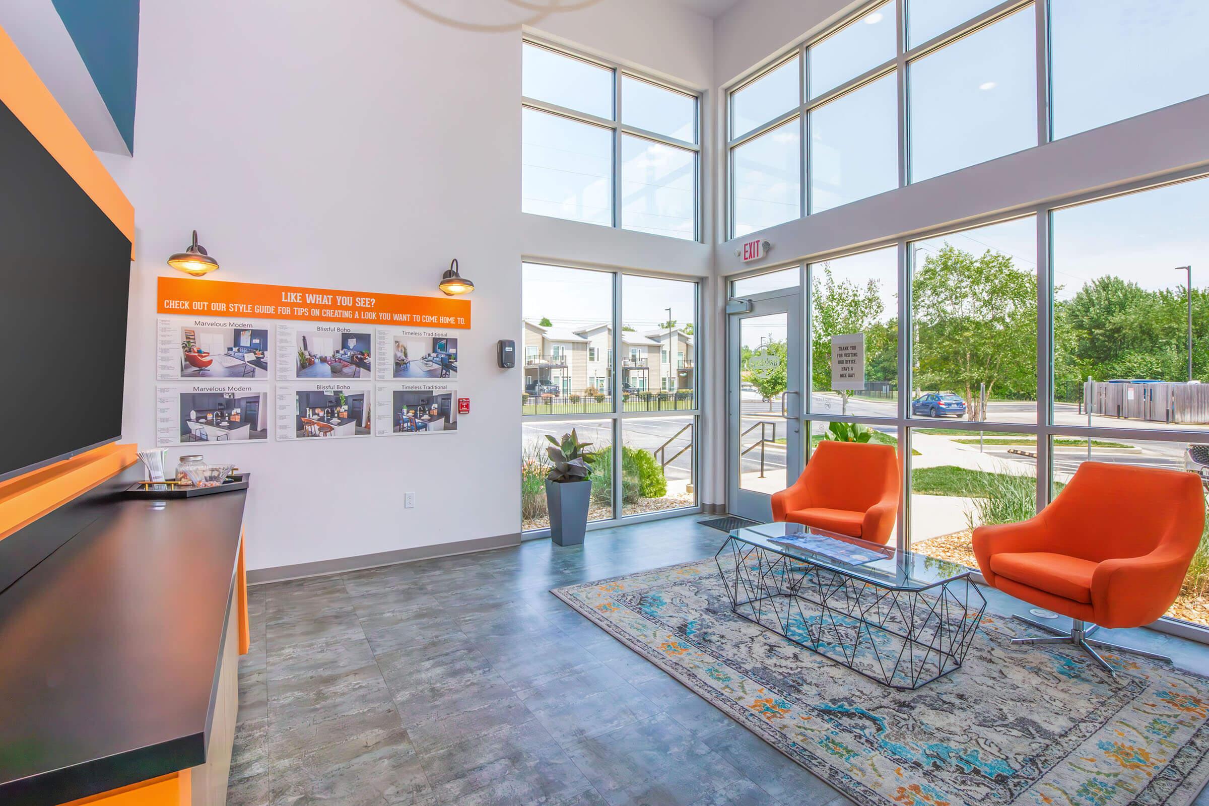 Bright and modern interior of a reception area featuring large windows, two orange chairs, a coffee table, and a decorative rug. A wall displays information with images, and there are potted plants. Natural light fills the space, creating a welcoming atmosphere.