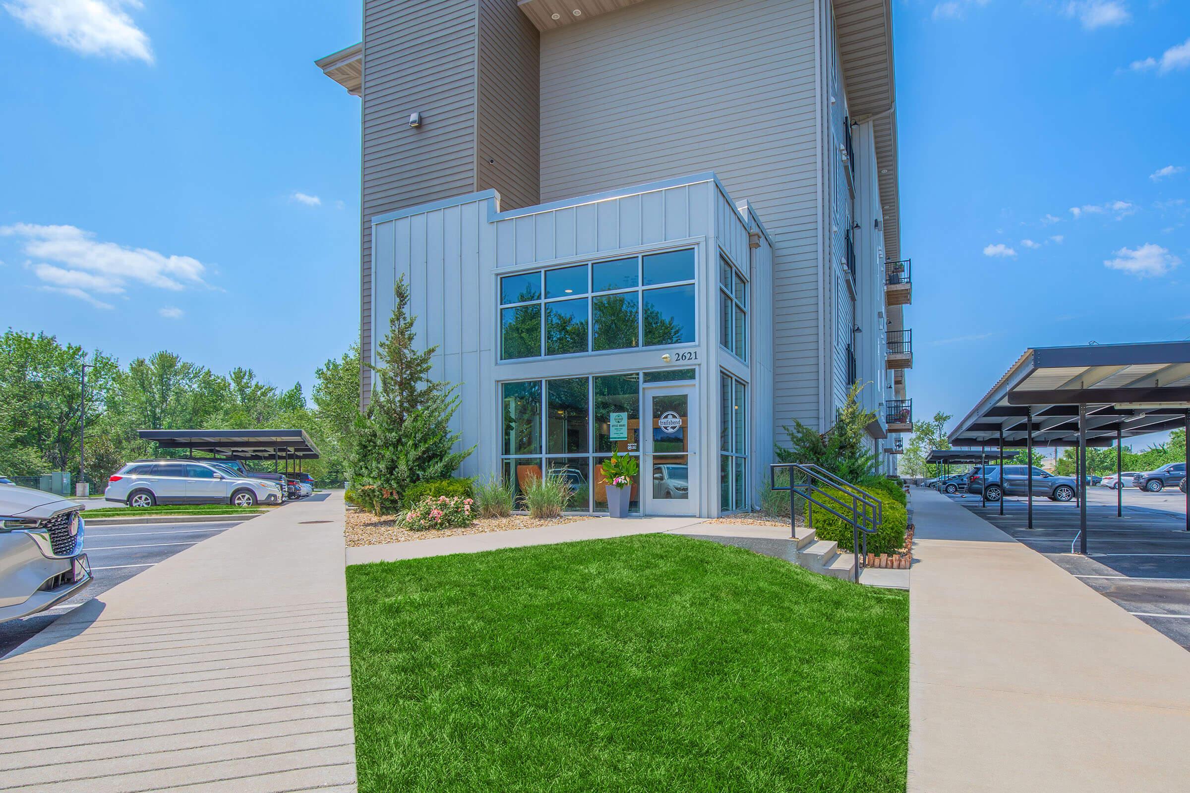 Modern building entrance with large glass windows, surrounded by lush green grass and landscaped shrubs. A paved pathway leads to the entrance, and there are parked cars in a shaded area nearby. The sky is clear with a few clouds, indicating a sunny day.