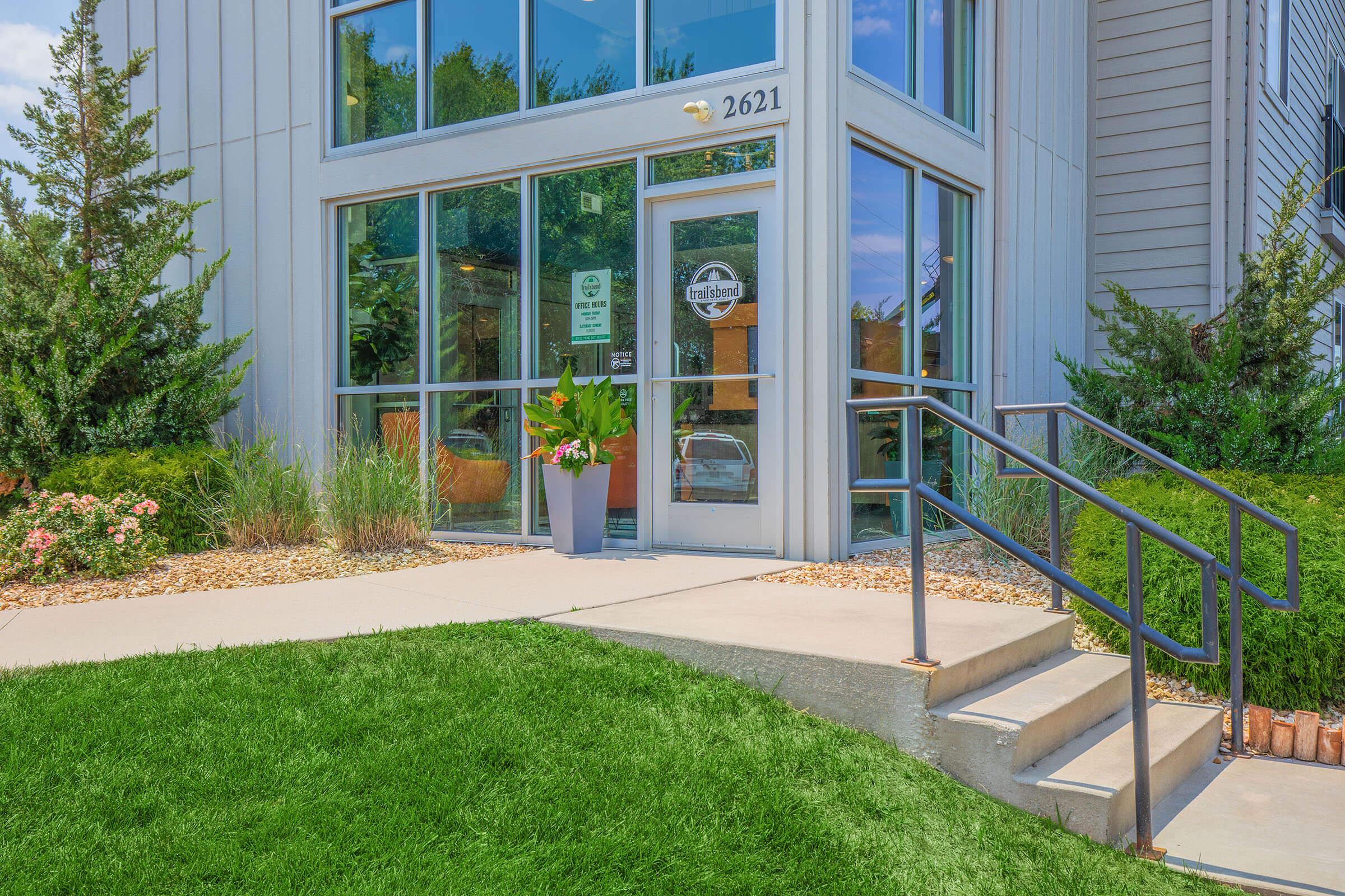 A modern building entrance with large glass windows. A door labeled "2621" is flanked by decorative plants in a planter. Concrete steps with a railing lead up to the entrance, surrounded by well-maintained grass and flower beds. Green shrubs add to the landscaping.
