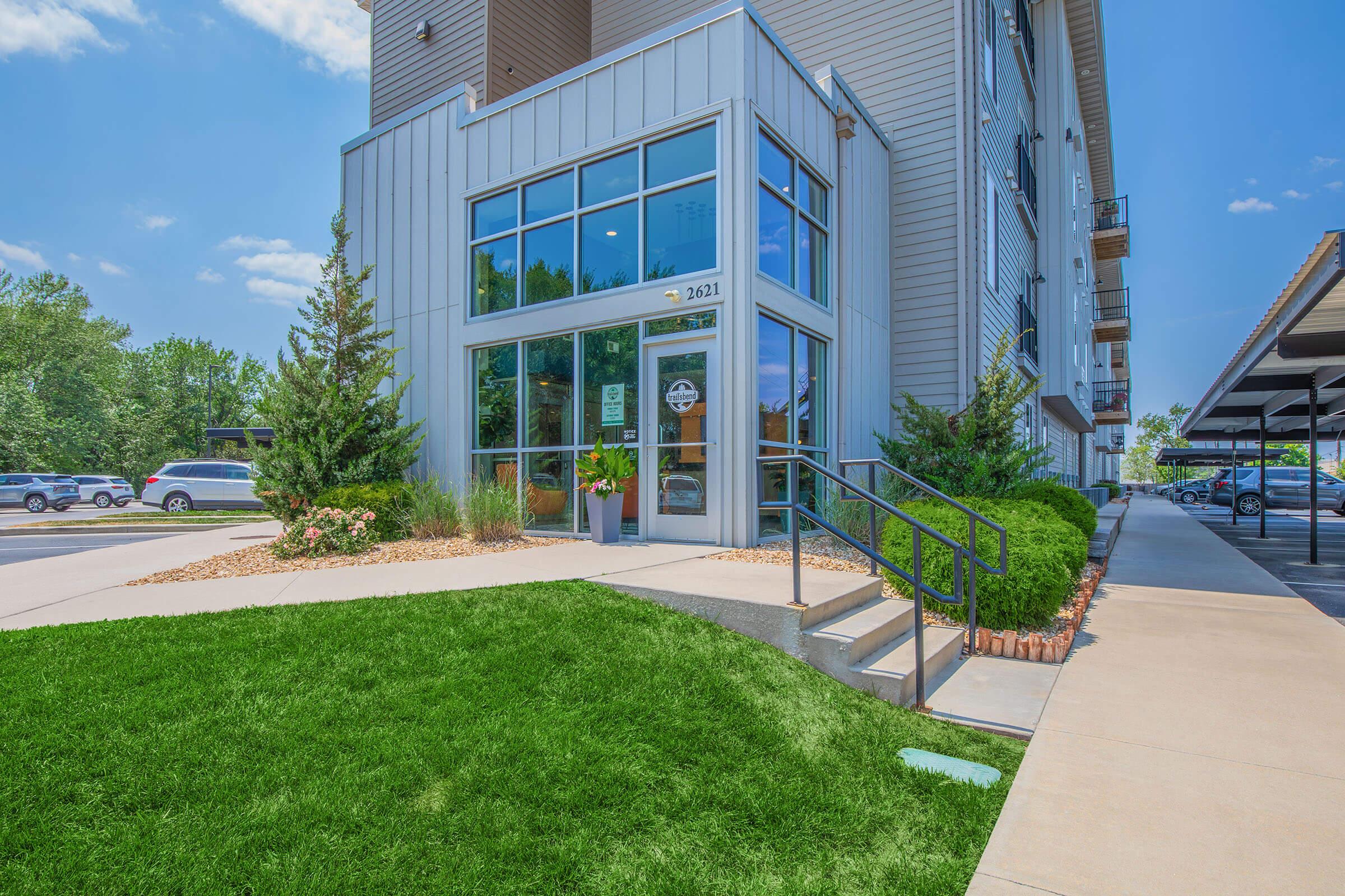 Modern building entrance with large glass doors, surrounded by neatly landscaped greenery. A pathway leads up to the entrance, which features decorative plants and shrubs. The exterior has a mix of wood and metal finishes, with a parking area visible in the background under a sunny blue sky.
