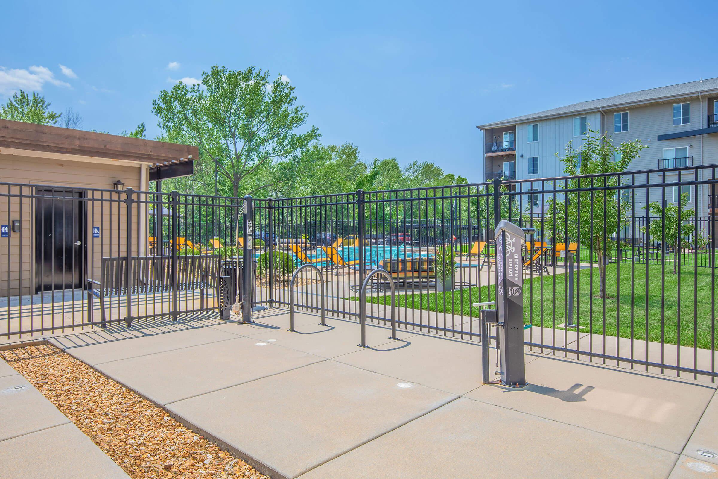 A fenced outdoor pool area with lounge chairs, surrounded by greenery. There’s a building in the background and a pathway leading to the gate. The sky is clear and blue, creating a bright, inviting atmosphere.