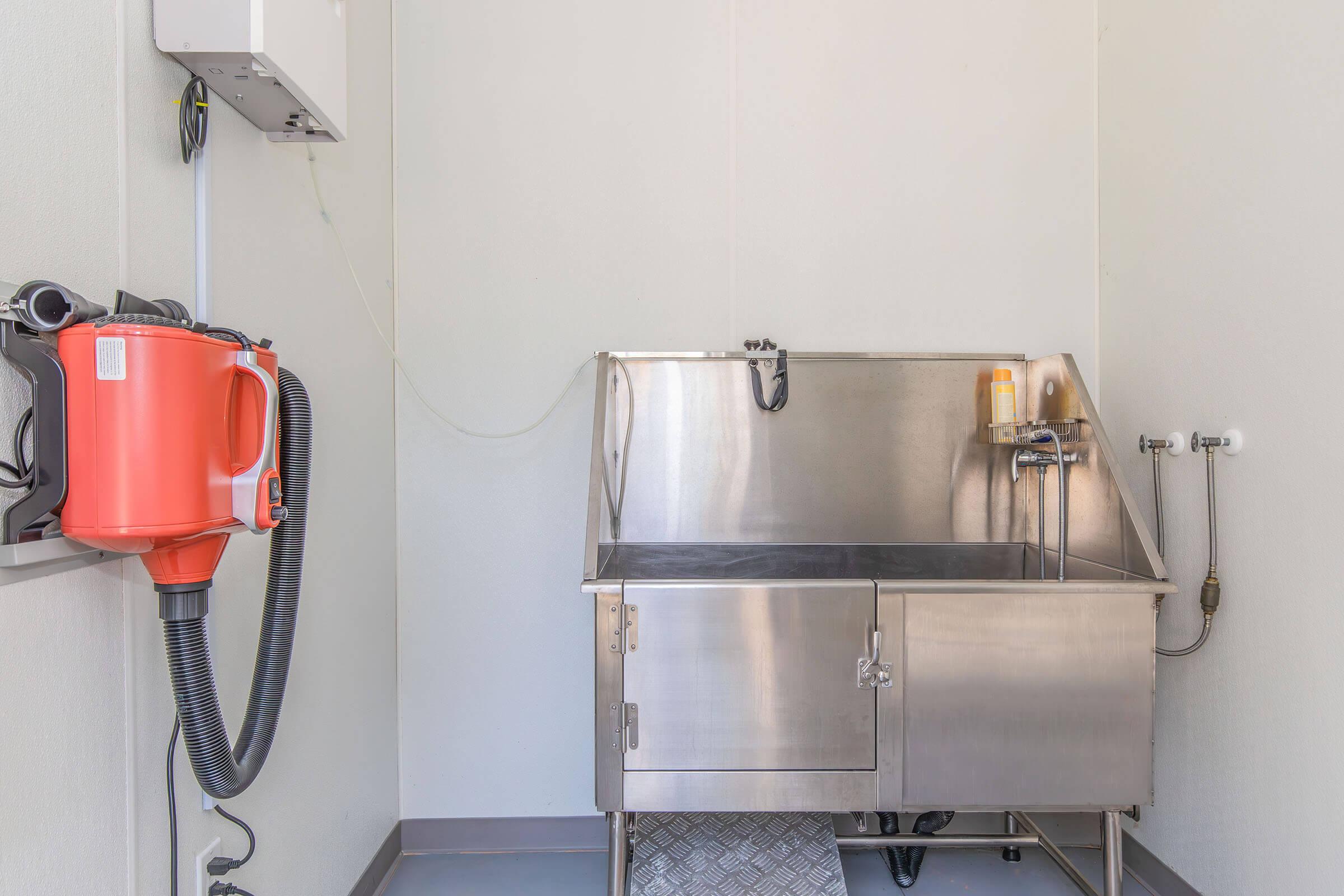 A clean, stainless steel utility sink is shown in a well-lit room. The sink features faucets and a drain, with a shelf above it holding an orange vacuum cleaner attached to a wall. The walls are light-colored and there's a minimalistic design, emphasizing functionality.