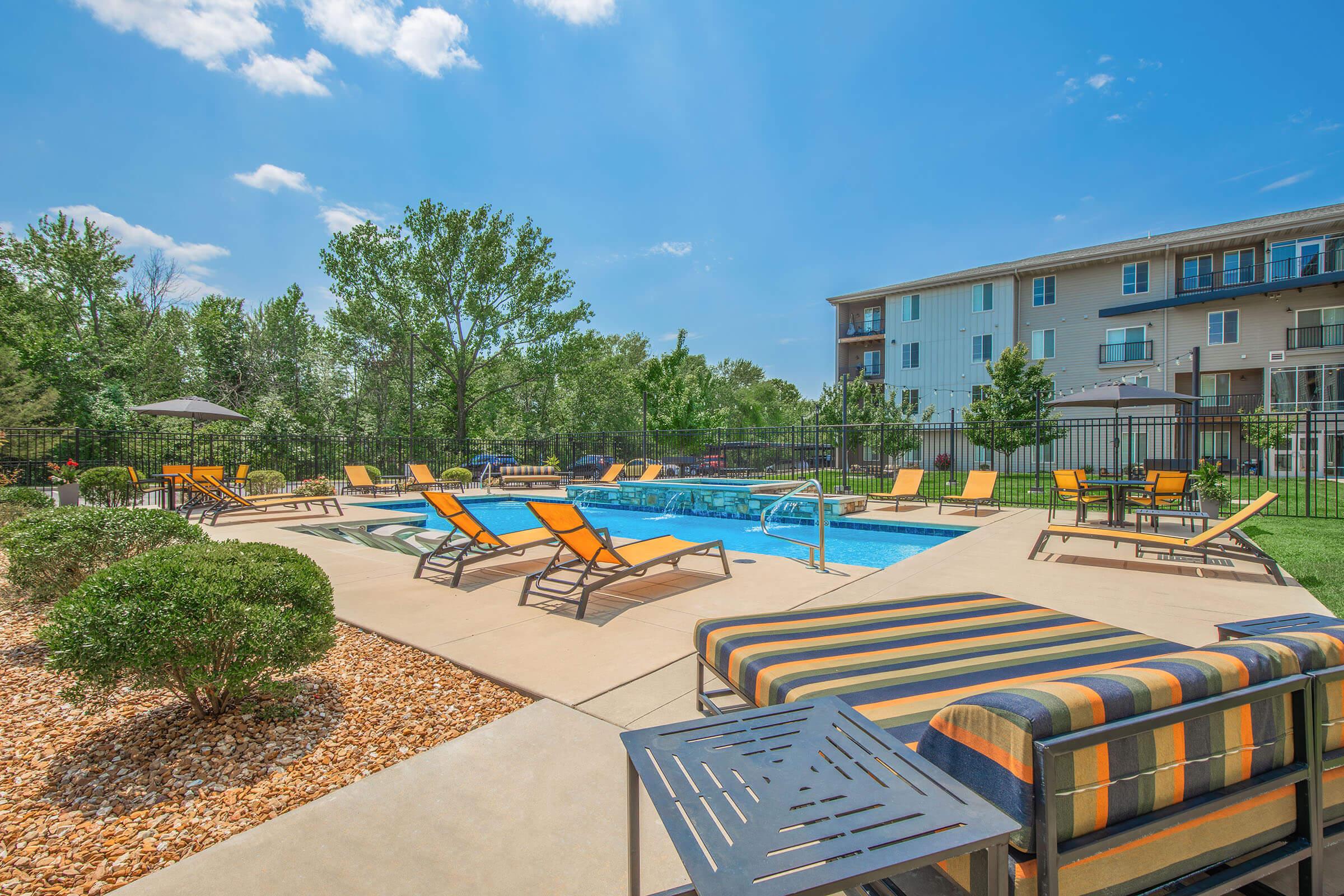 A serene outdoor pool area with lounge chairs and umbrellas surrounded by greenery. The clear blue sky and bright sun enhance the inviting atmosphere. In the background, an apartment building can be seen, creating a cozy residential setting.
