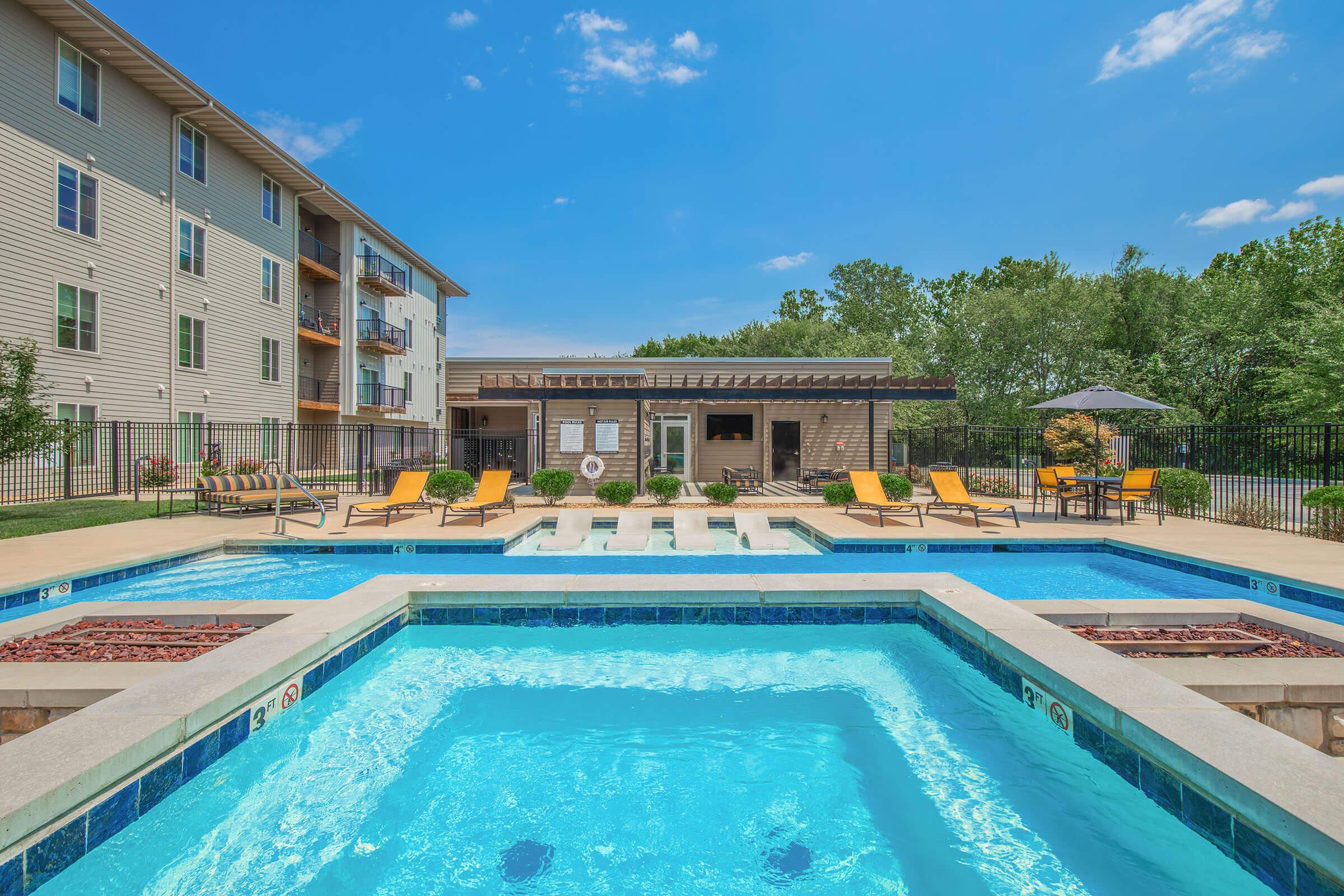 A sunny outdoor pool area with several blue swimming pools, surrounded by lounge chairs. In the background, a modern building with balconies and a shaded seating area. Lush greenery and a clear blue sky create a relaxing atmosphere.