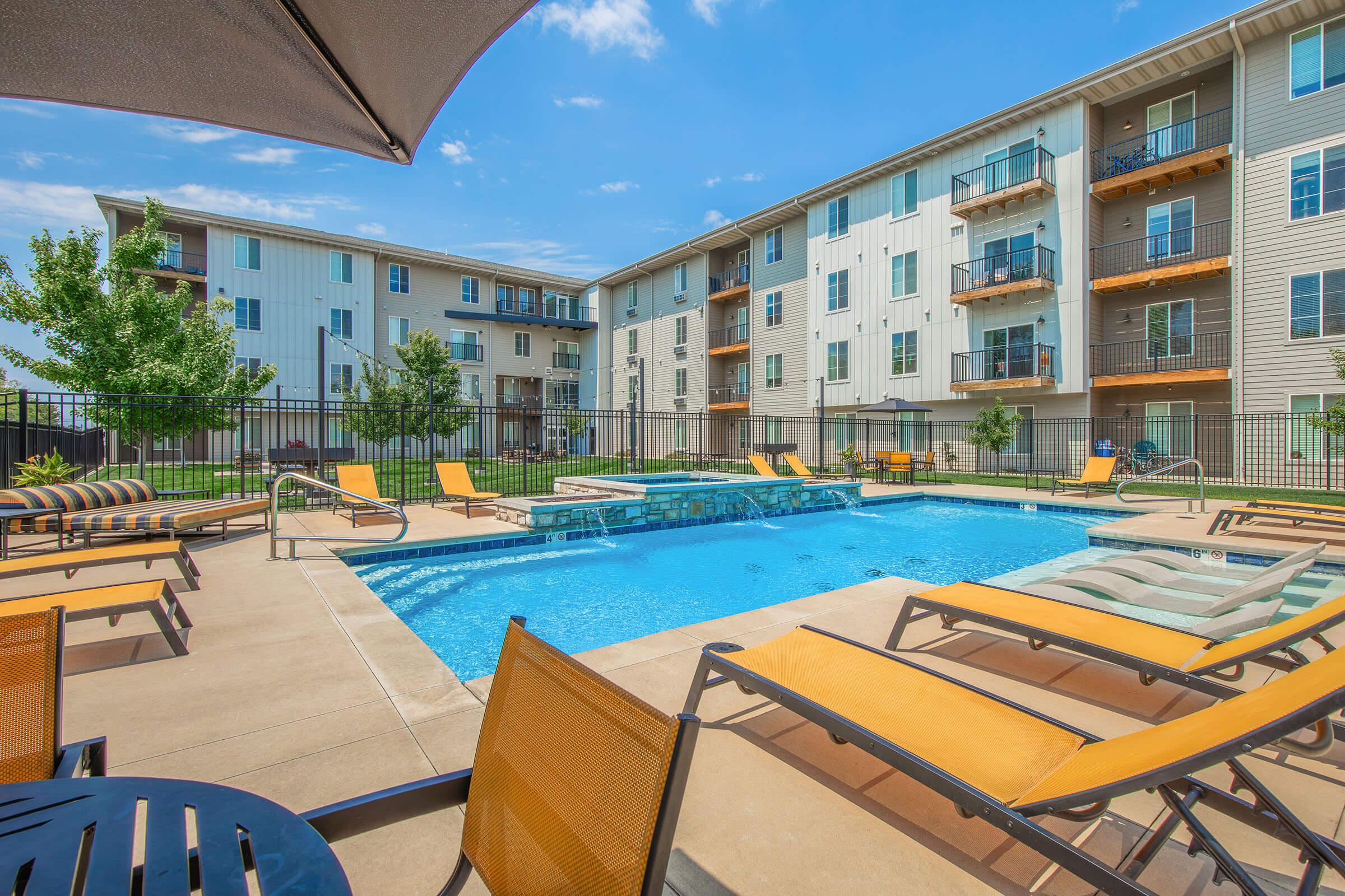 A sunny outdoor swimming pool surrounded by lounge chairs, with trees and apartment buildings in the background. The pool area is enclosed with a fence, featuring a bright blue sky and inviting atmosphere.