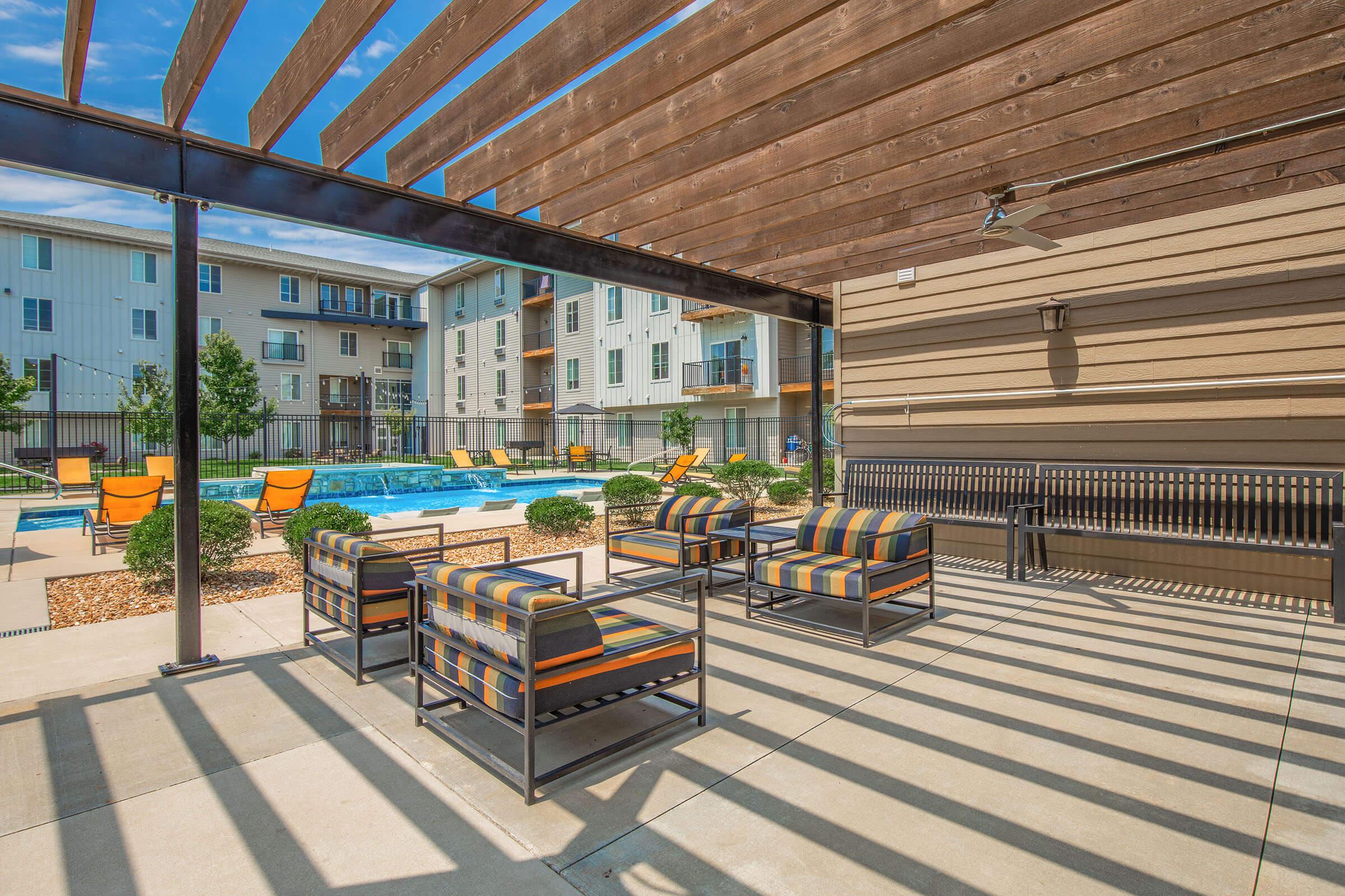 Outdoor lounge area featuring striped chairs and a coffee table under a wooden pergola. In the background, there's a swimming pool surrounded by landscaped greenery and sun loungers. The scene is bright and inviting, with modern apartment buildings visible in the distance.