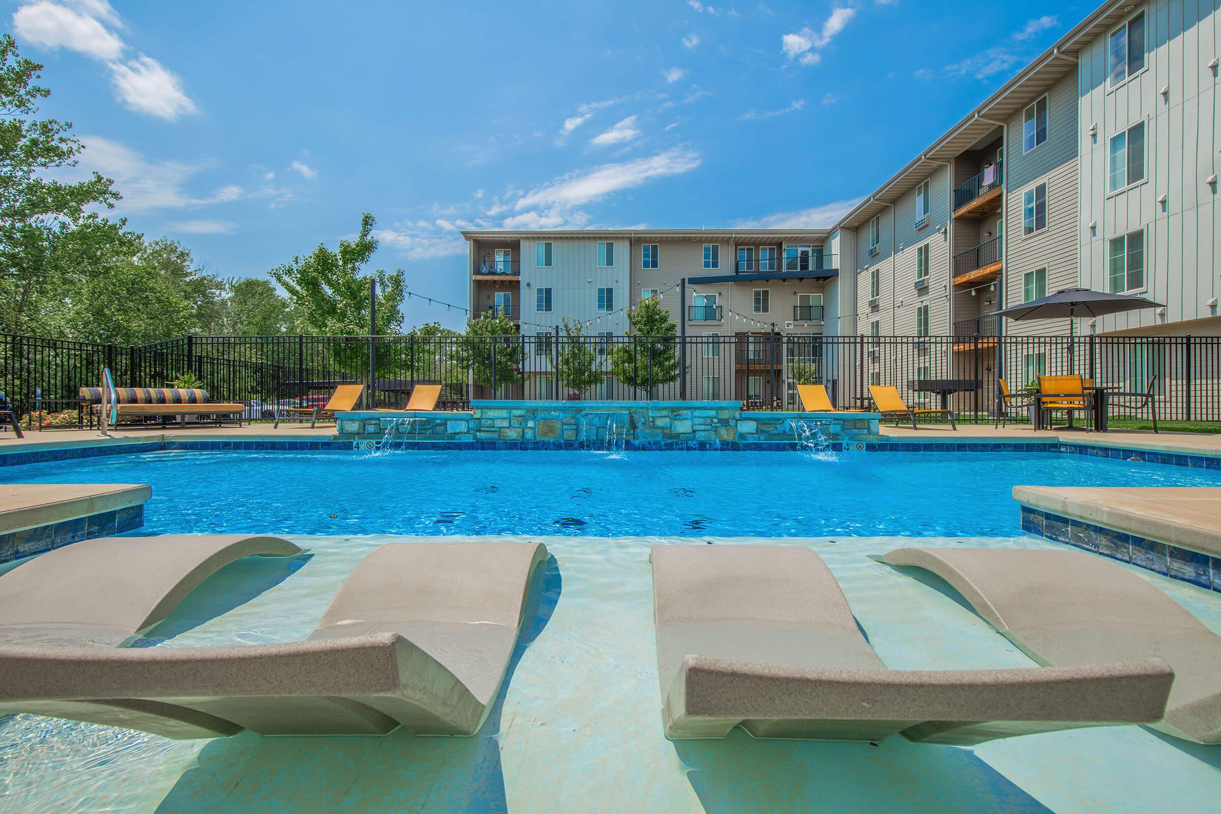 A clear blue swimming pool with two sun loungers partially submerged at the edge. In the background, a modern apartment building is visible along with green trees. The sky is bright with a few scattered clouds, and poolside chairs are arranged around the area, creating a relaxing outdoor atmosphere.