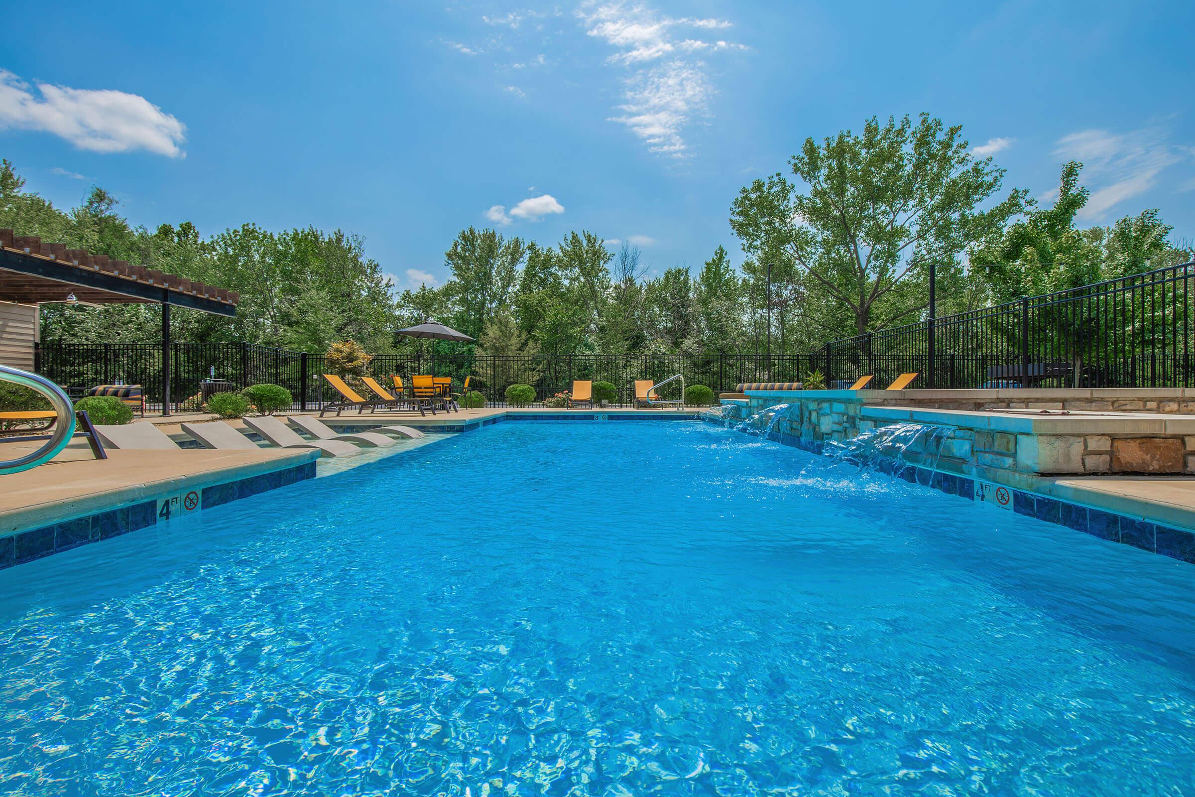 A clear blue swimming pool surrounded by lounge chairs and leafy greenery. There are umbrellas for shade, and a stone wall features a water feature that cascades into the pool under a bright blue sky with fluffy white clouds. The setting appears inviting and relaxing, ideal for a summer day.
