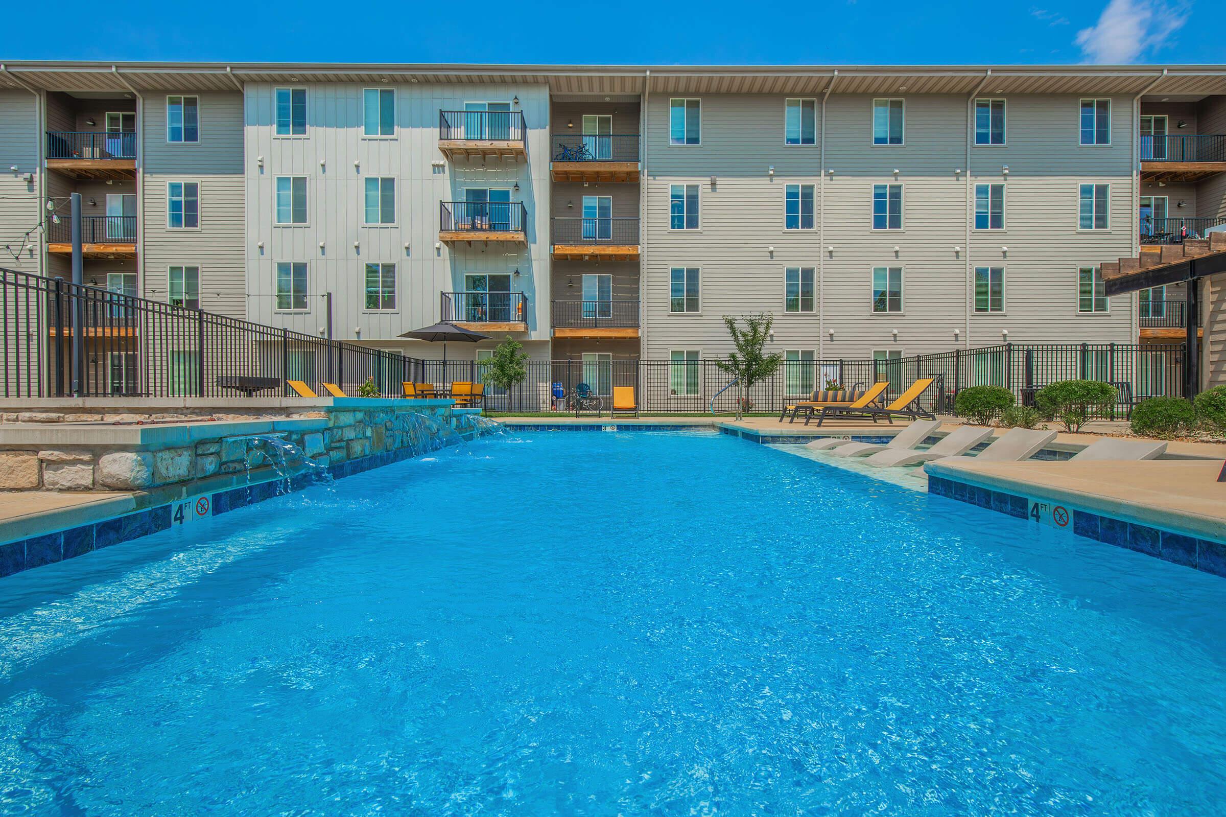 A clear blue swimming pool surrounded by a manicured area, with lounge chairs and umbrellas. In the background, a three-story apartment building features balconies and multiple windows. The sky is bright and partly cloudy, creating a sunny atmosphere.