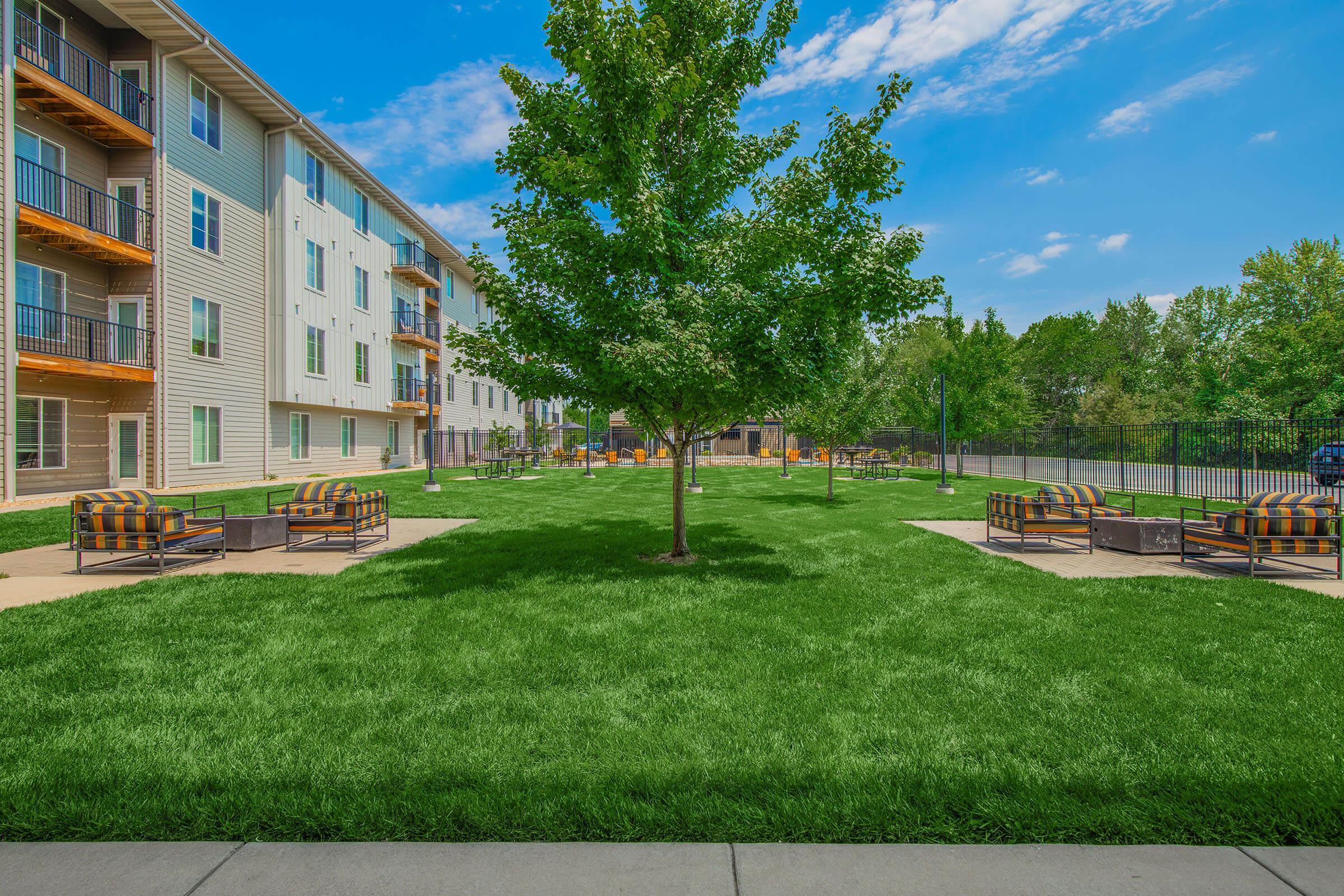 A well-maintained outdoor space featuring a lush green lawn, a tree at the center, and several seating areas with comfortable chairs. Surrounding the lawn are modern apartment buildings and a clear blue sky, creating a welcoming and relaxing atmosphere.