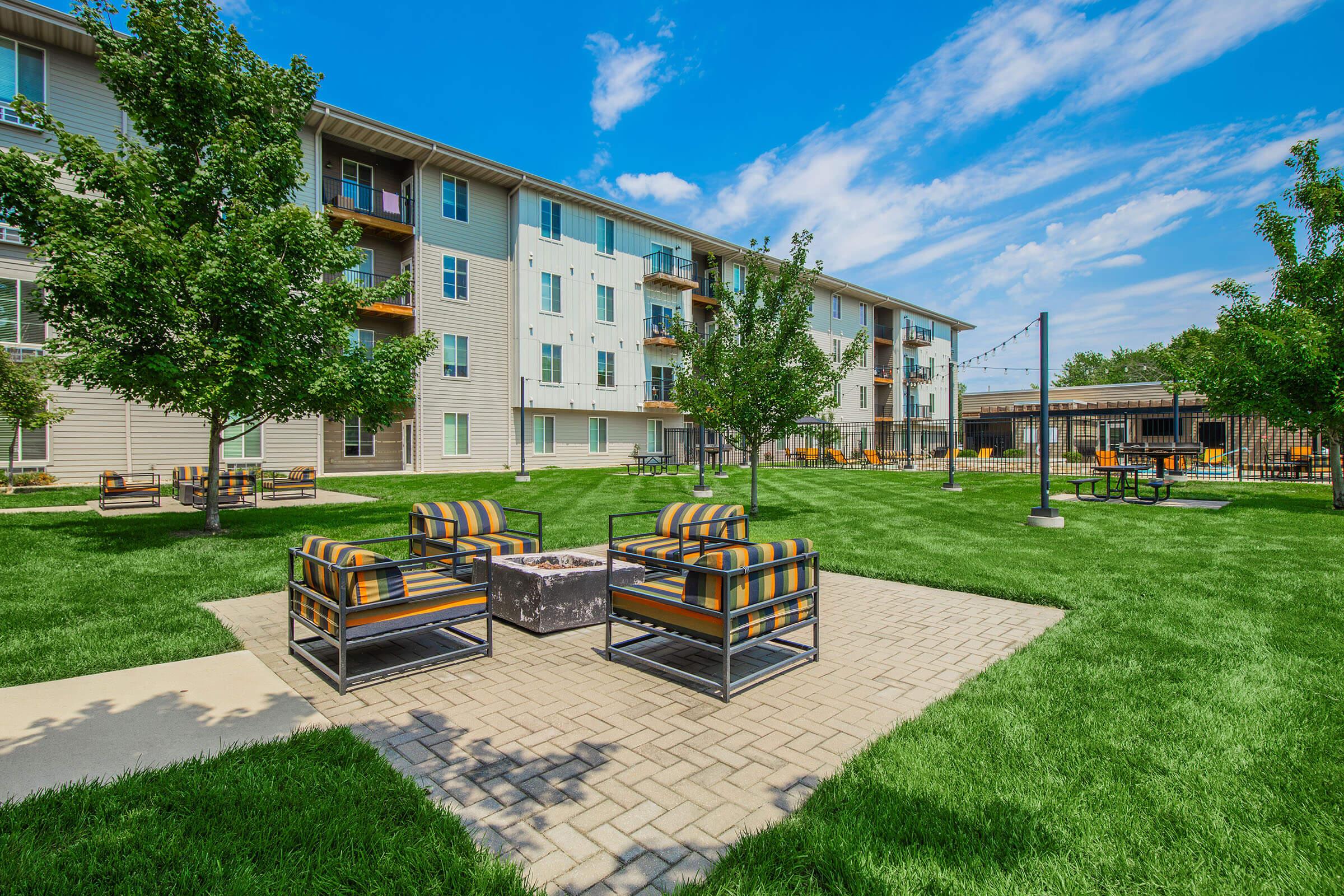A spacious outdoor area featuring a well-manicured lawn, colorful seating arrangements, and a fire pit. Surrounding the seating are young trees, and in the background, a multi-story apartment building is visible under a bright blue sky with scattered clouds, creating a welcoming community atmosphere.