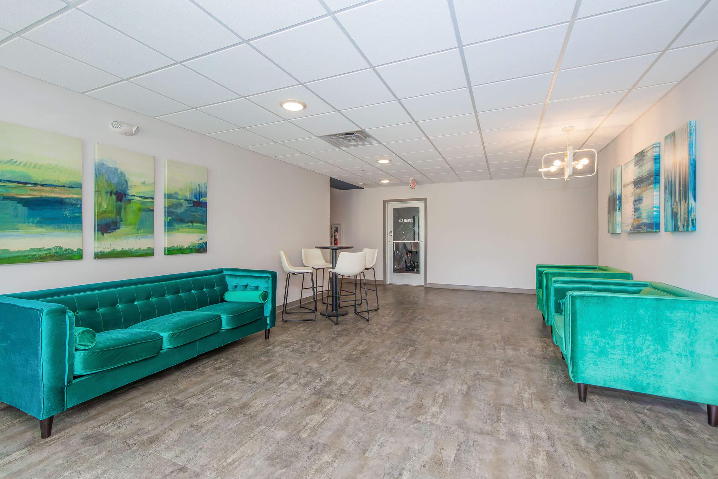 A modern lounge area featuring two bright teal sofas and matching chairs. The space is adorned with abstract art pieces in green and blue tones on the walls. A small dining table with high stools is positioned in the center, under a stylish light fixture, contributing to a contemporary ambiance.