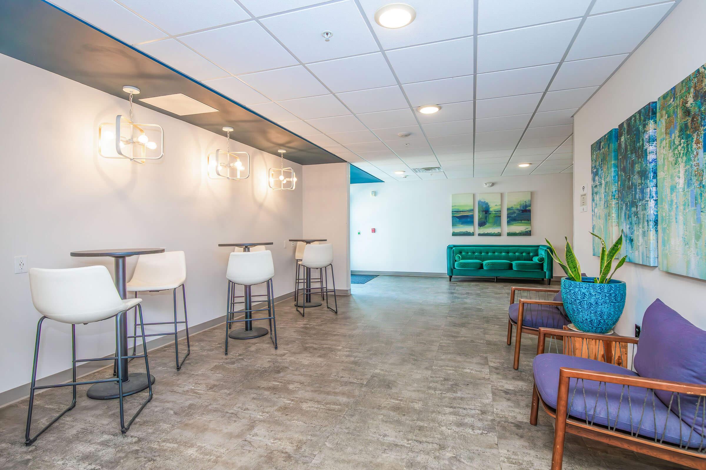 A modern, well-lit hallway featuring two tall tables with stools, a green couch, and decorative artwork on the walls. There are potted plants and a mix of seating options, creating a welcoming atmosphere in a contemporary space.