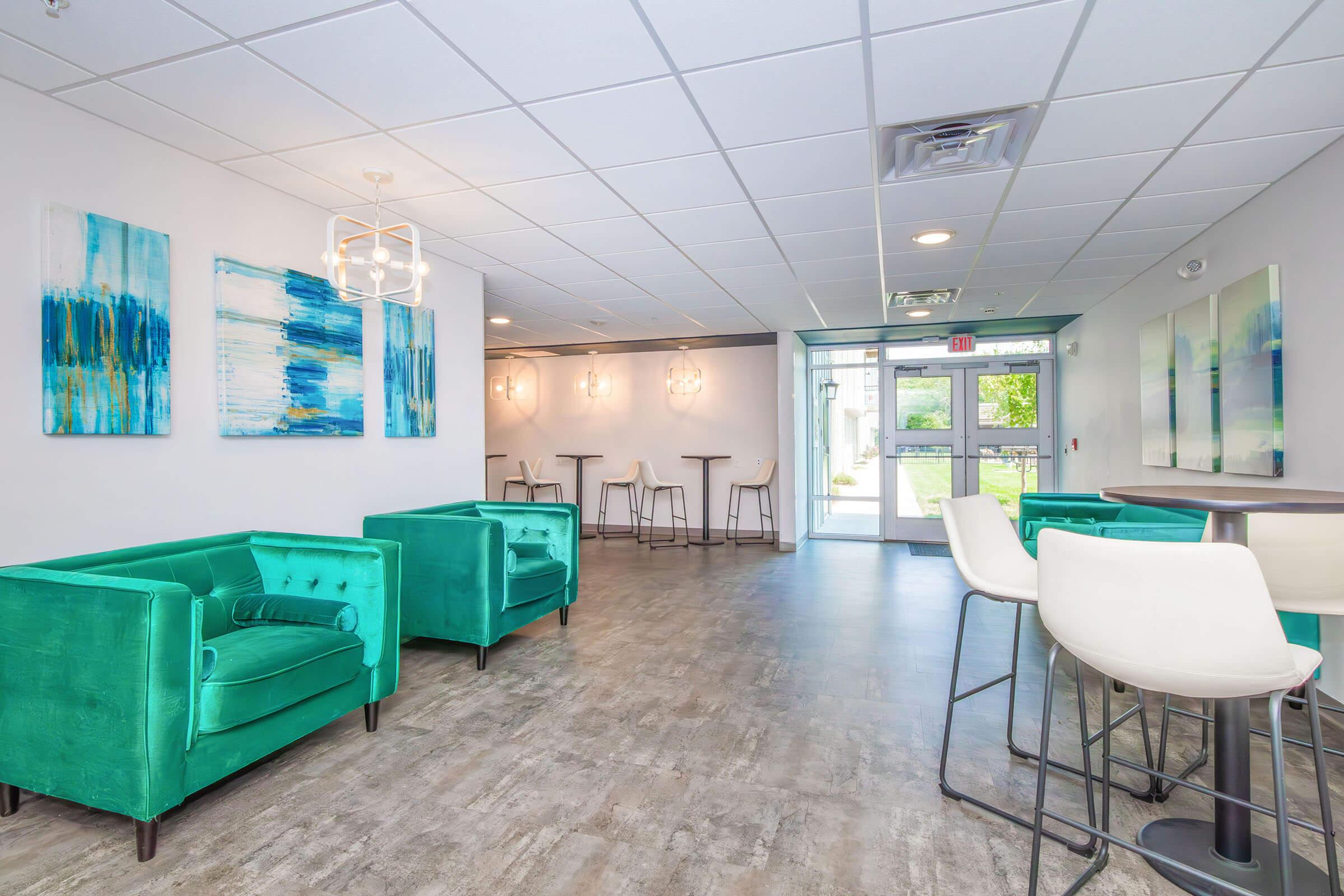 A modern, brightly lit lounge area featuring two teal sofas and high-top tables. The walls are adorned with abstract blue and white artwork. Large windows allow natural light, and the entrance is visible in the background, creating an inviting atmosphere.