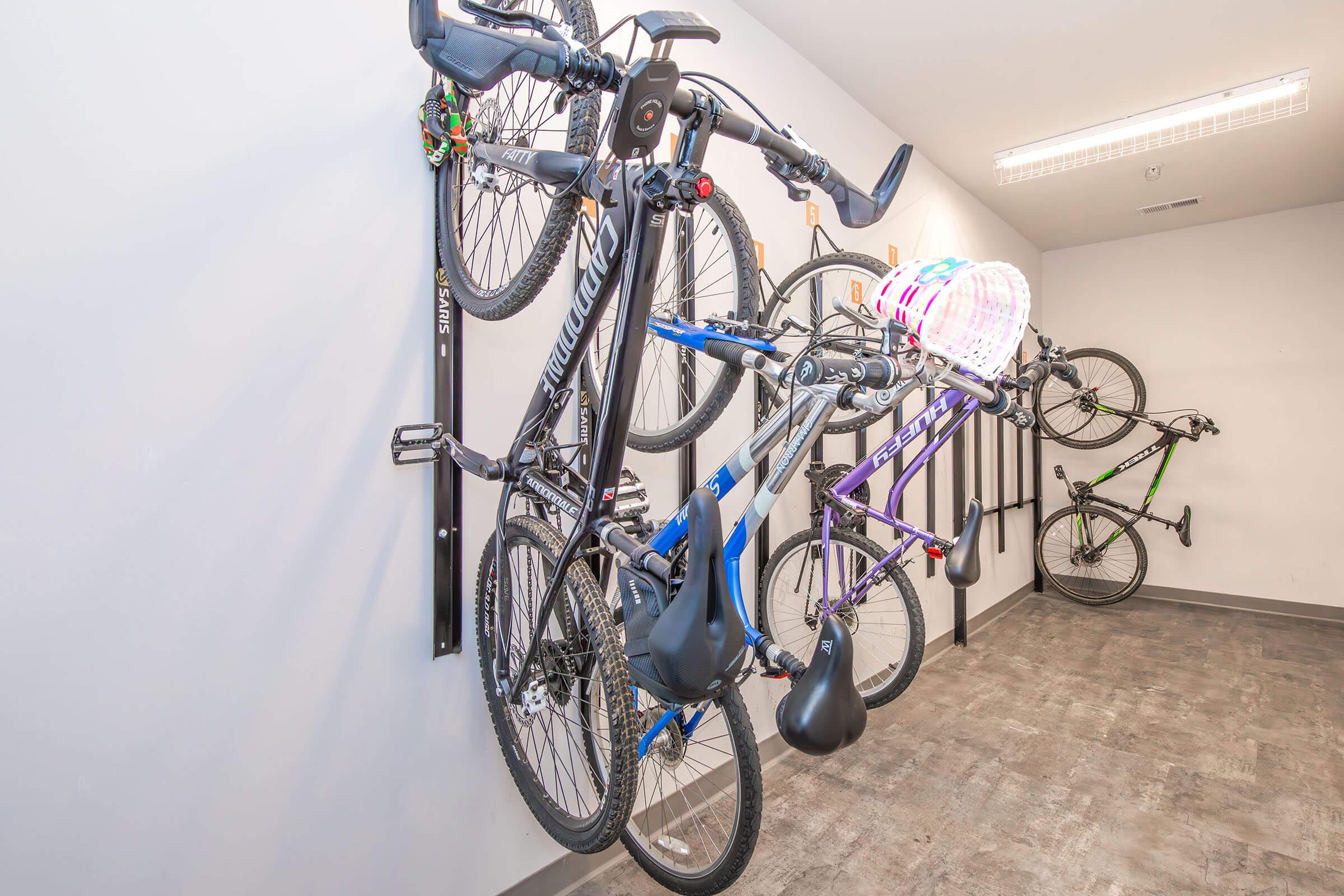 A well-organized bike storage area featuring multiple bicycles mounted on wall racks. The bikes are of various colors and styles, with a colorful helmet hanging on one. The background is a clean, light-colored wall and a gray floor, creating a neat and functional space for storing bikes.