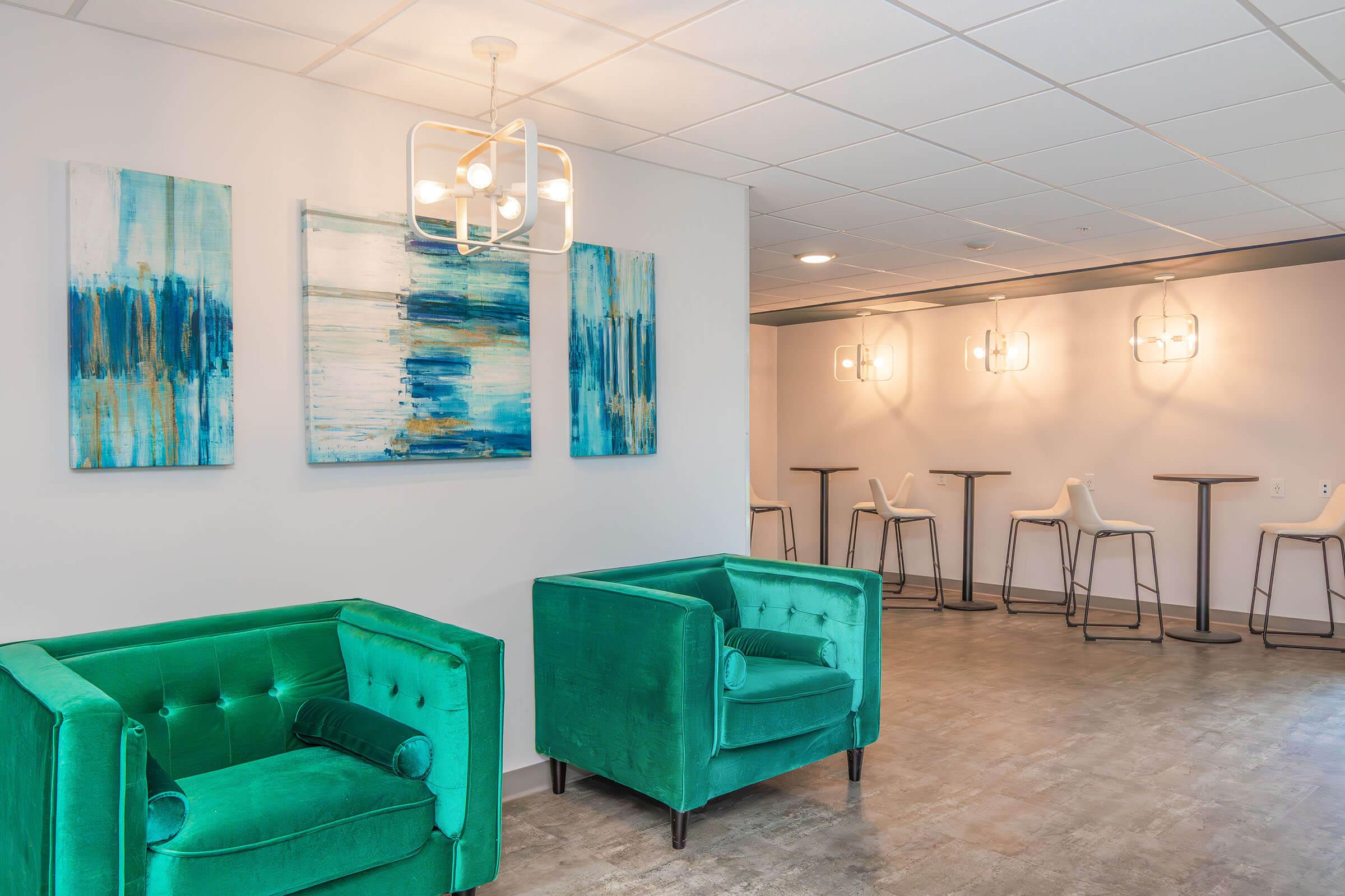 A modern lounge area featuring two green velvet armchairs, abstract artworks in blue and white on the wall, and bar-height tables with stools in the background. The space is well-lit with a stylish light fixture and has a contemporary, inviting atmosphere.