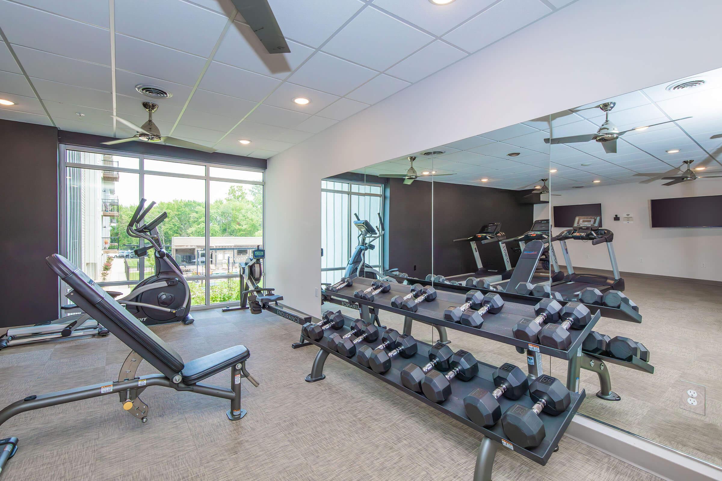 A modern fitness room featuring a variety of exercise equipment. There are dumbbells arranged on a rack, an adjustable bench, and treadmills positioned next to large windows that allow natural light. The room is well-lit with ceiling fans and a mirror on one wall.