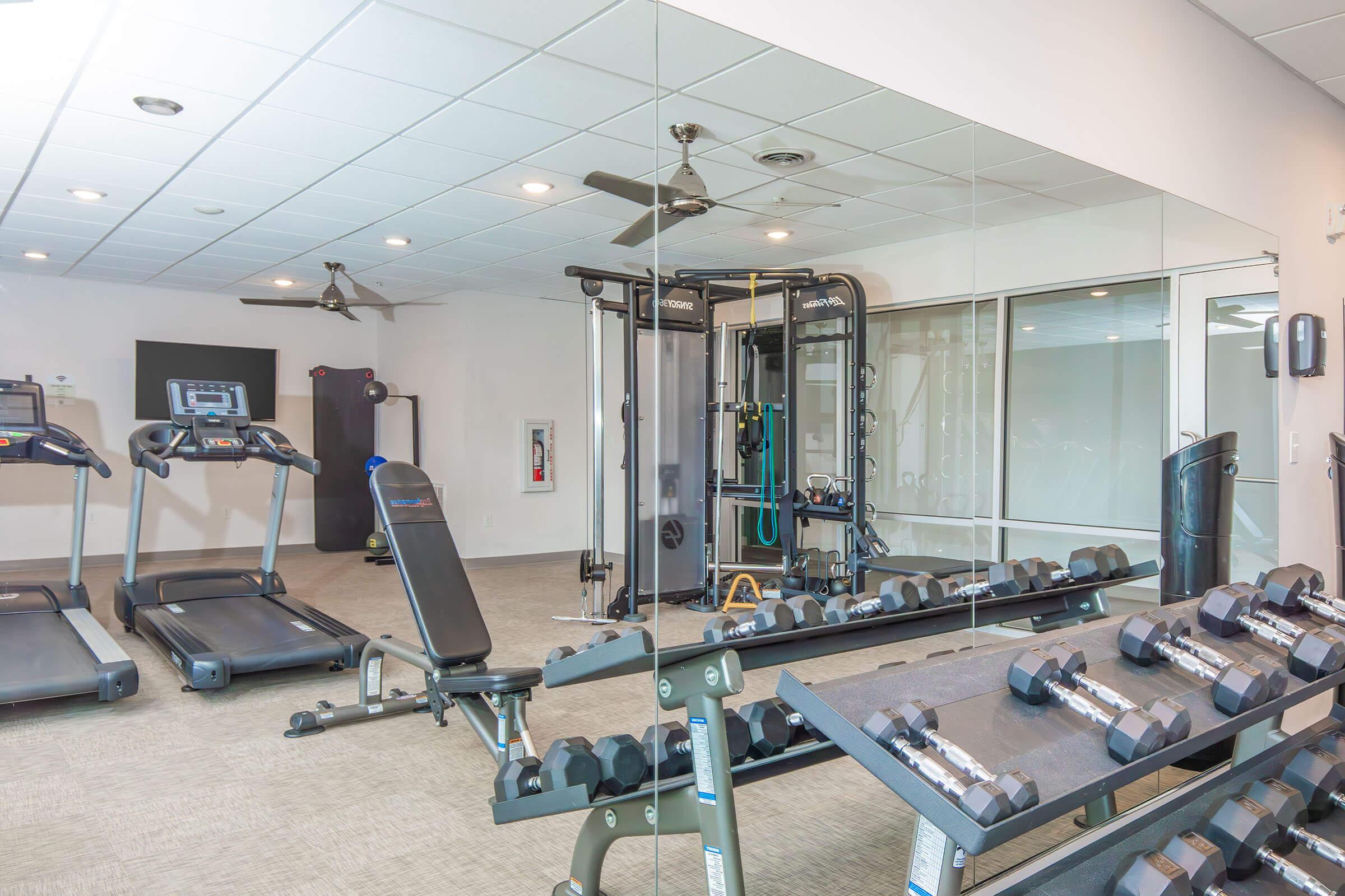 A modern gym interior featuring several fitness machines, including two treadmills, a multi-station weight machine, and a bench. A rack of dumbbells is prominently displayed in the foreground, with large mirrors on one wall reflecting the equipment and room's design. Bright lighting creates an inviting atmosphere.