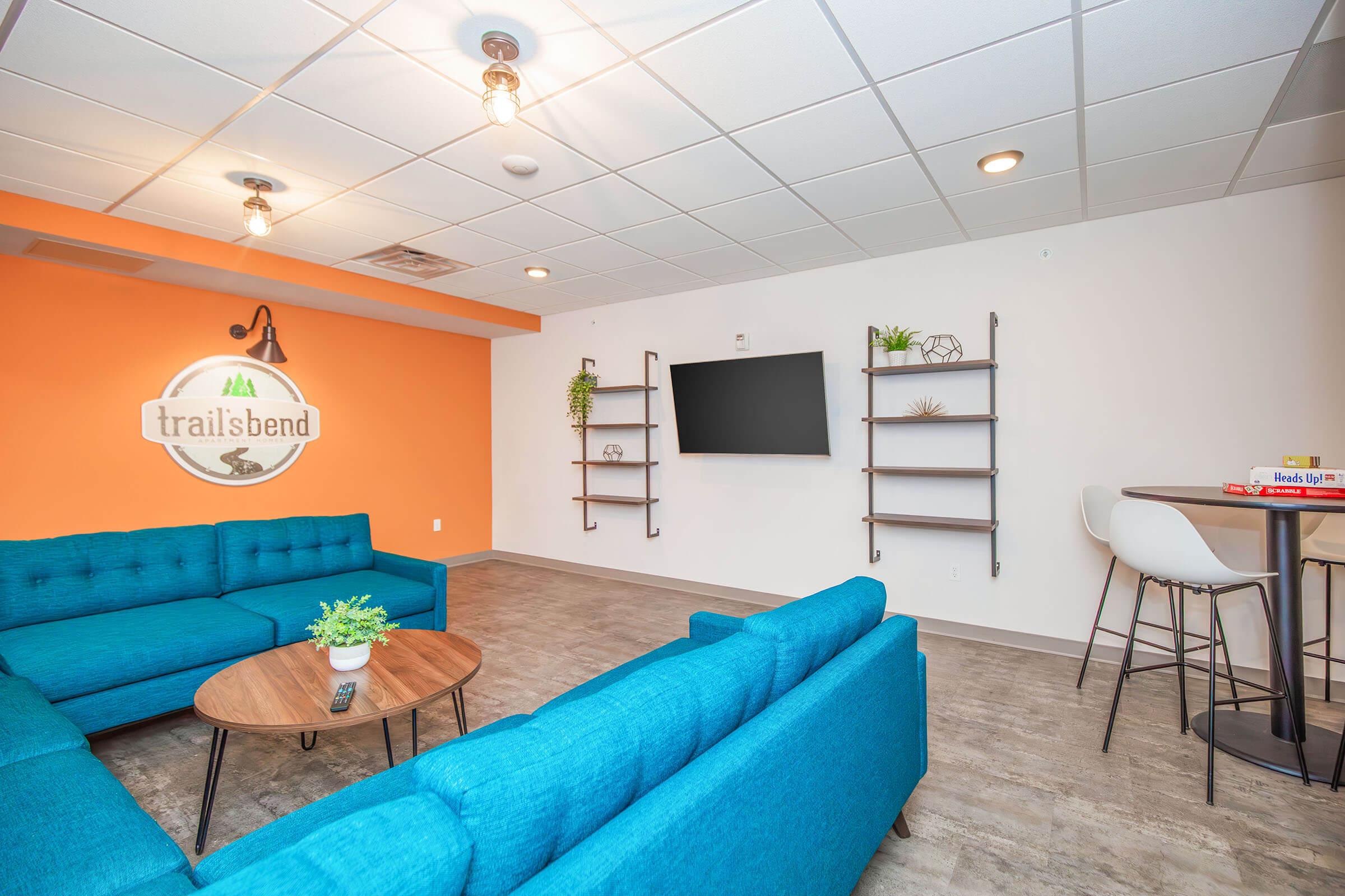 A modern lounge area featuring a bright orange accent wall and a circular sign saying "trailsbend." There's a blue sectional sofa around a round wooden coffee table, along with shelves on the wall displaying plants and decorative items. A flat-screen TV is mounted on the wall, and there are bar stools at a small table.