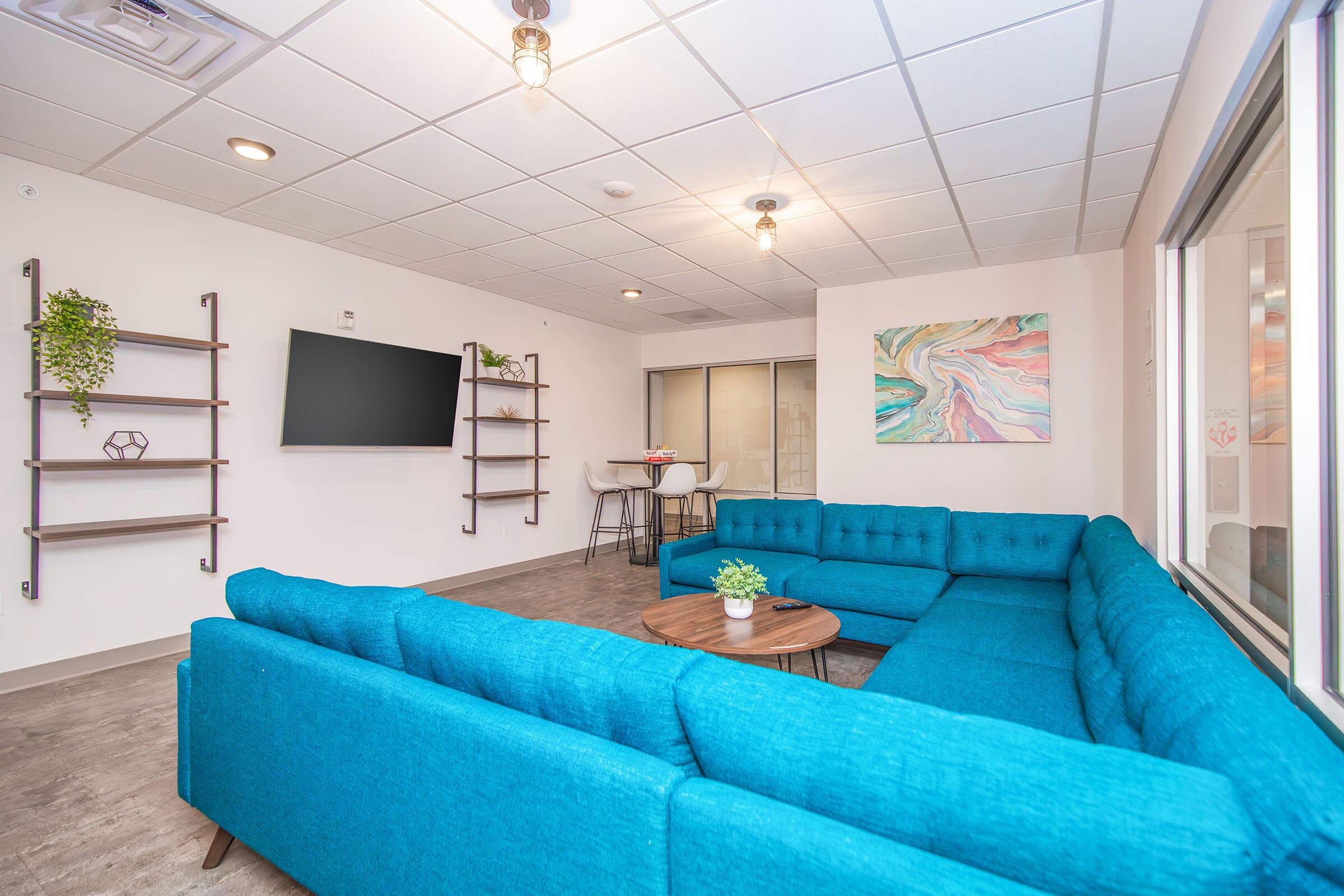 Modern communal lounge with a large turquoise sectional sofa, a round wooden coffee table, and two wall-mounted shelves. A flat-screen TV is mounted on the wall, and there’s a small dining area with white stools. Decor includes a colorful abstract painting and potted plants. Bright and inviting atmosphere.