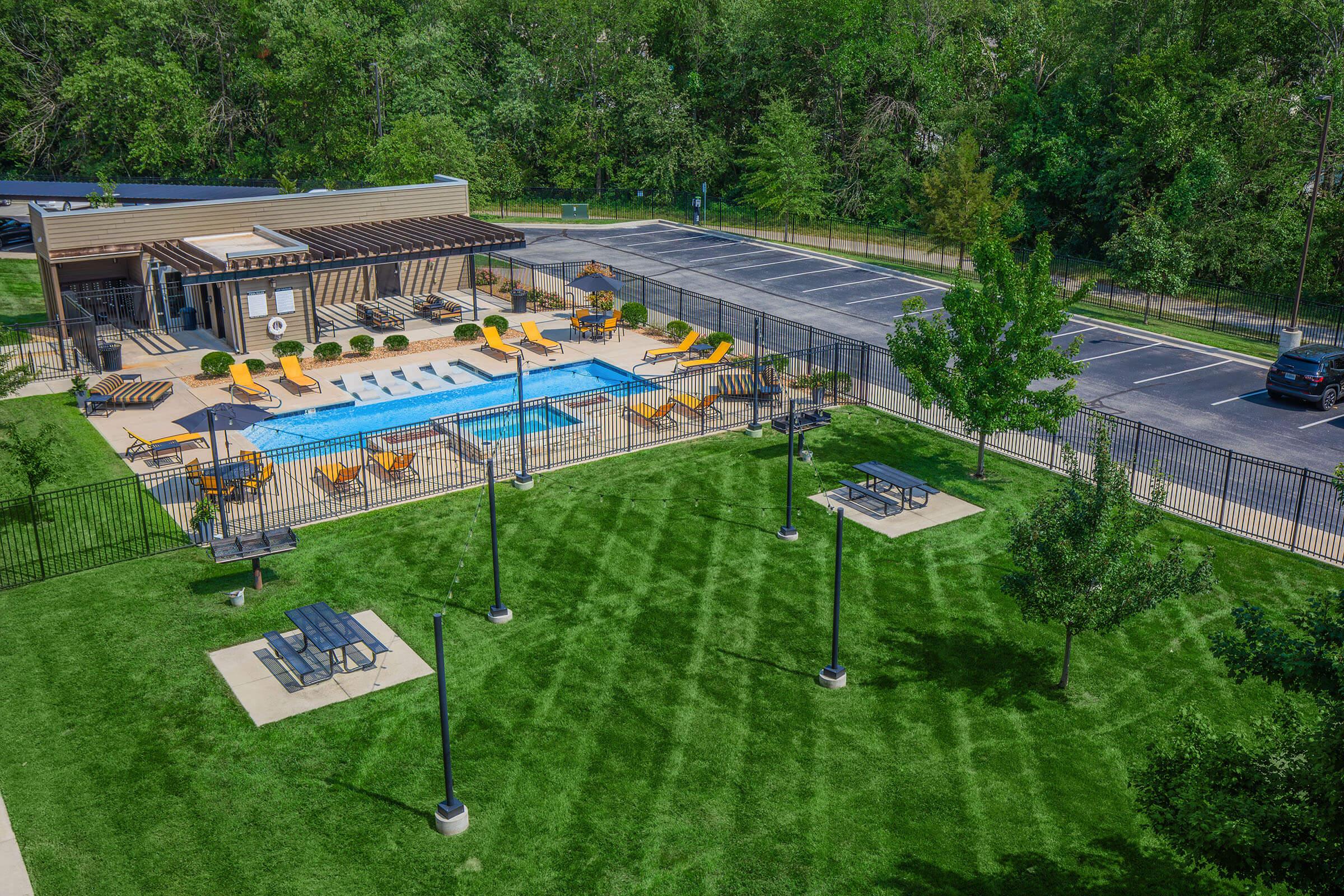 Aerial view of a landscaped area featuring a swimming pool surrounded by lounge chairs and umbrellas. Nearby, there are picnic tables and trees. The area is fenced, and a parking lot is visible in the background, with several cars parked. Sunlight casts shadows across the manicured grass.