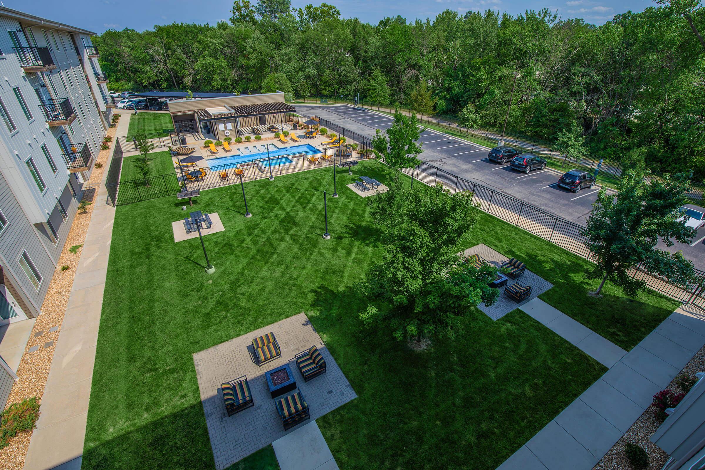 Aerial view of a landscaped outdoor area featuring a swimming pool surrounded by lounge chairs, wooden pergolas, and seating areas with fire pits. Lush green grass and trees provide a natural backdrop. Nearby, a parking area is visible along a road.