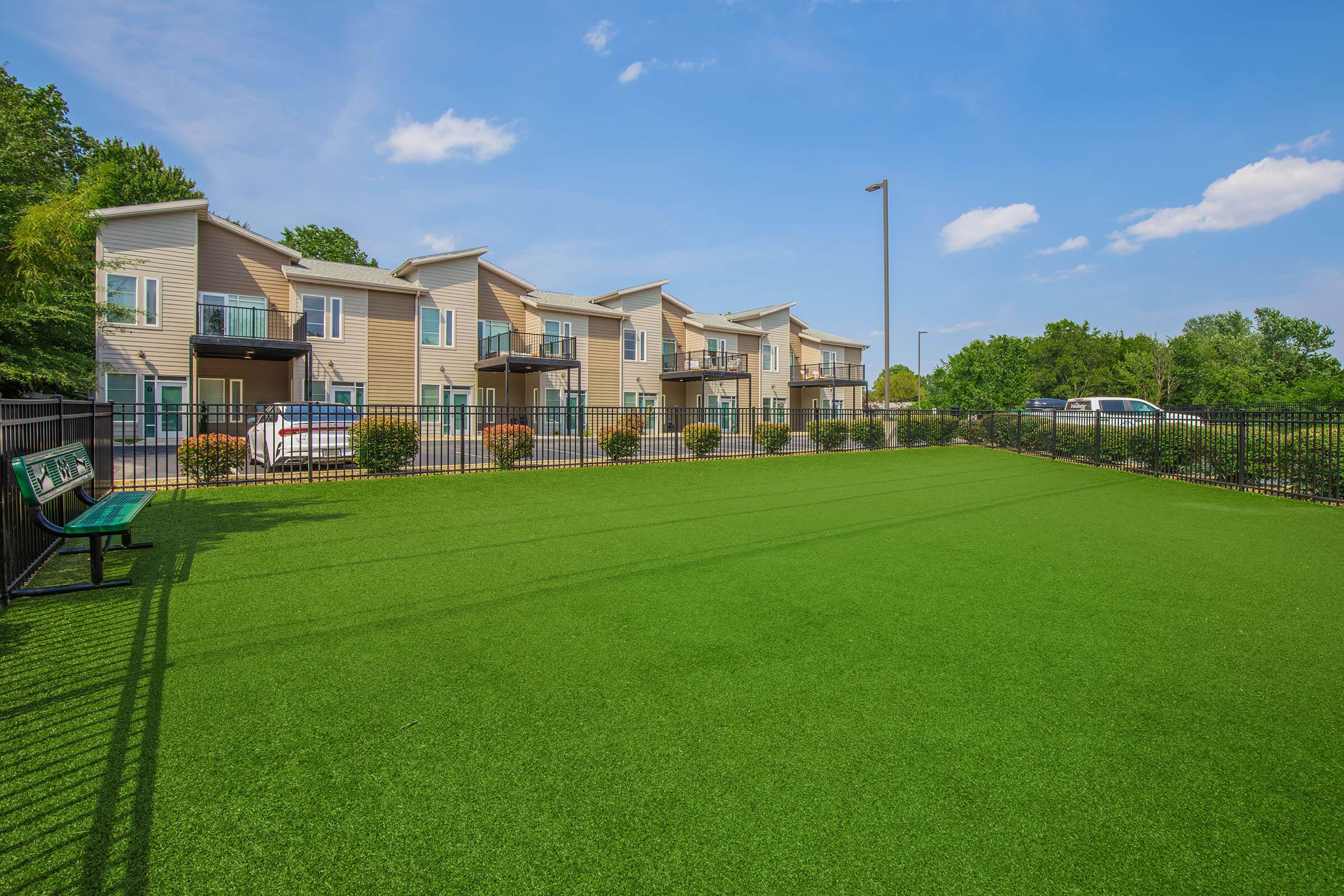 A well-maintained grassy area with artificial turf surrounded by a black metal fence. In the background, there are modern apartment buildings with balconies. Blue sky with a few clouds complements the serene setting, and parked cars are visible nearby.