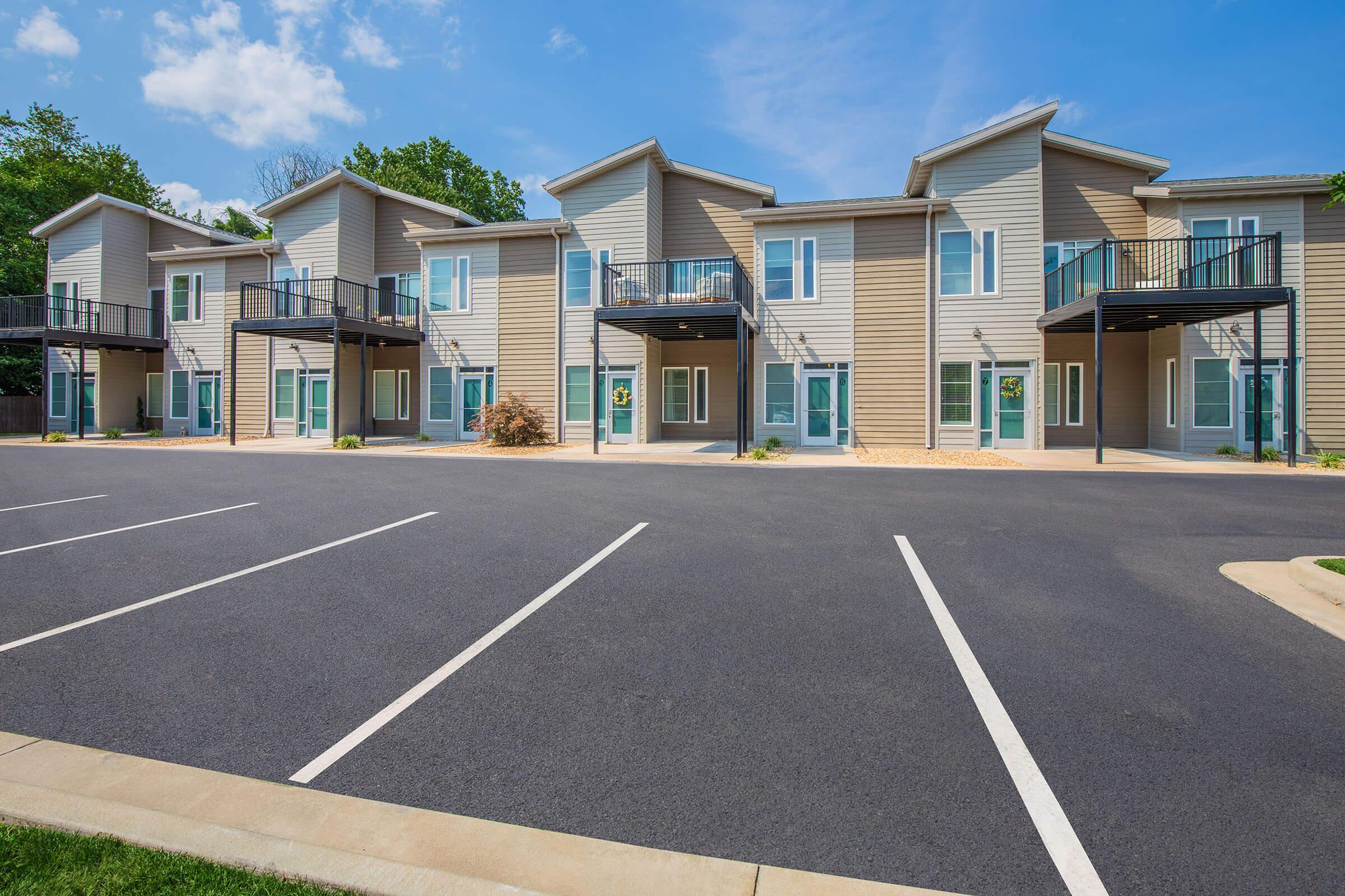 A modern apartment complex with multiple units featuring balconies, set against a bright blue sky. The landscape includes a paved parking area with marked spaces and neatly landscaped patches near the building, showcasing a clean and inviting exterior.