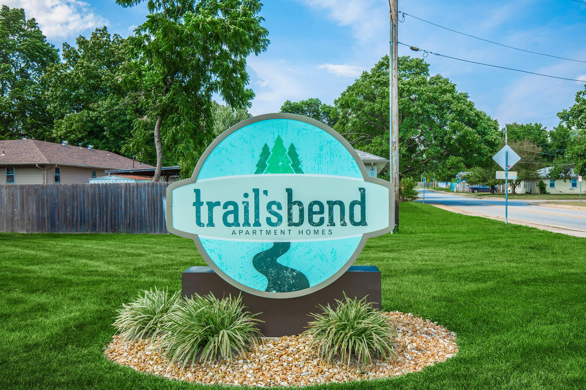 Sign for Trail's Bend Apartment Homes featuring a scenic design with a winding river and trees. The sign is surrounded by landscaped grass and decorative stones, with a clear blue sky in the background. Visible street and utility poles suggest a welcoming residential area.