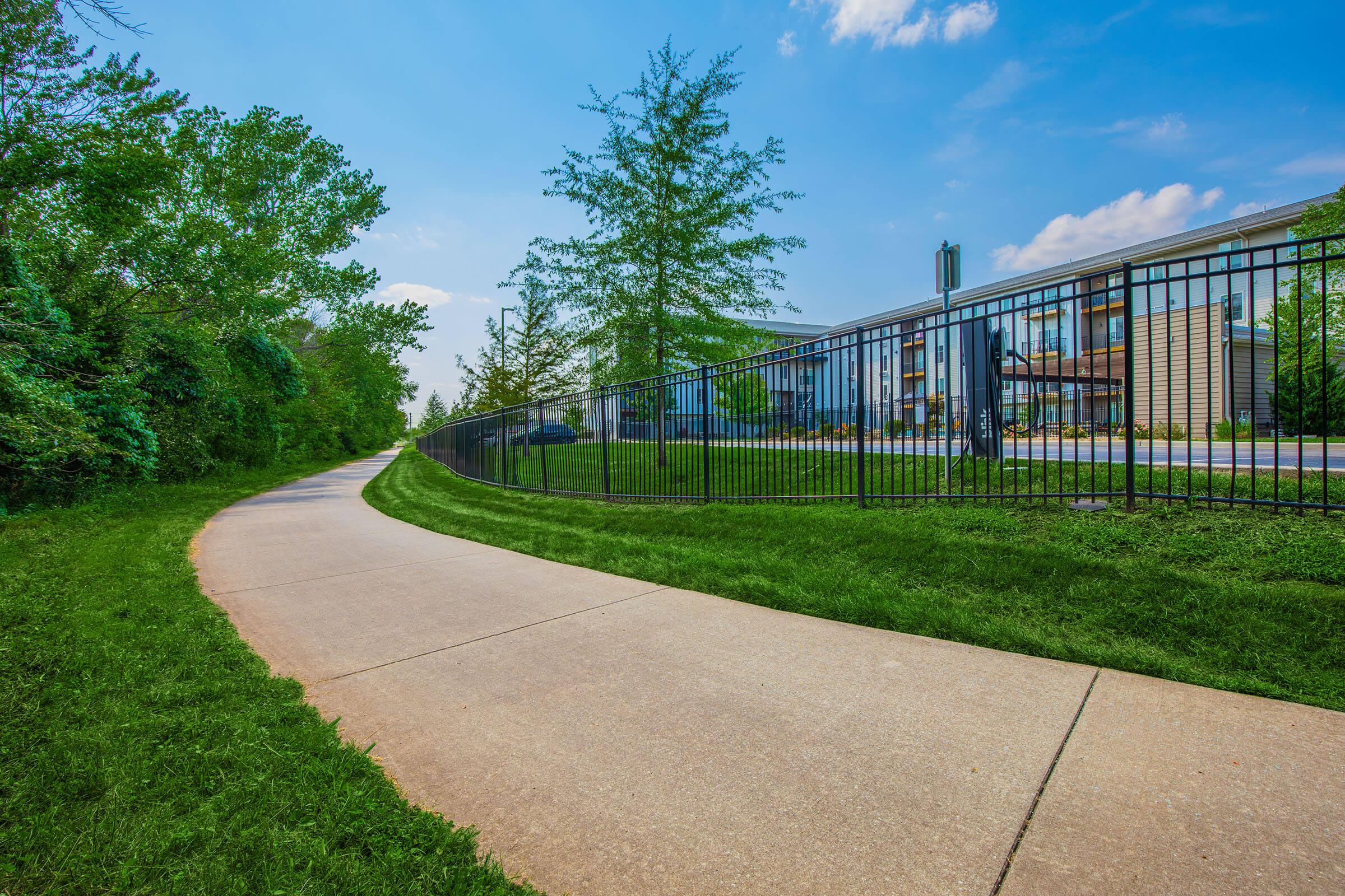 A paved path winds through a lush green area, bordered by trees and grass on one side. On the other side, a fence encloses a modern building with large windows, set against a bright blue sky with a few clouds. The scene conveys a sense of tranquility and nature.