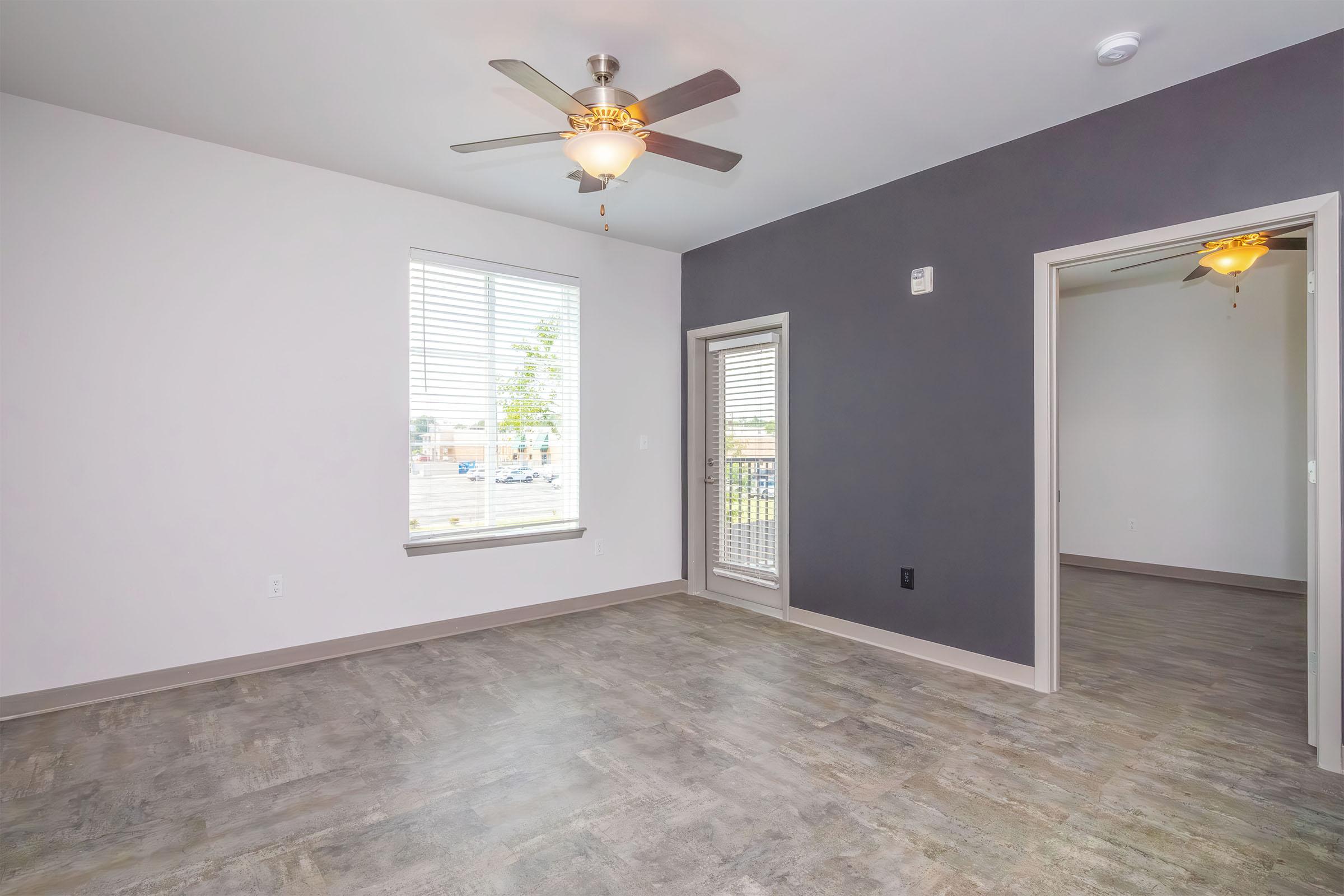 A spacious living room featuring light gray walls, hardwood-style flooring, and a ceiling fan. One wall is painted dark gray, and a large window with blinds allows natural light. A sliding door leads to a balcony. An adjacent room is visible through an open doorway.