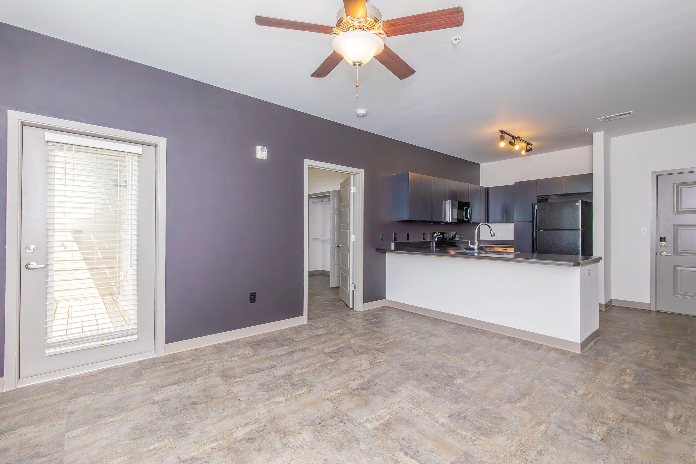 Spacious modern apartment with an open floor plan, featuring a kitchen area with dark cabinetry, a black refrigerator, and a ceiling fan. Large windows allow natural light, and there is a sliding door leading to a small balcony or patio. Neutral-toned walls complement the stylish flooring.