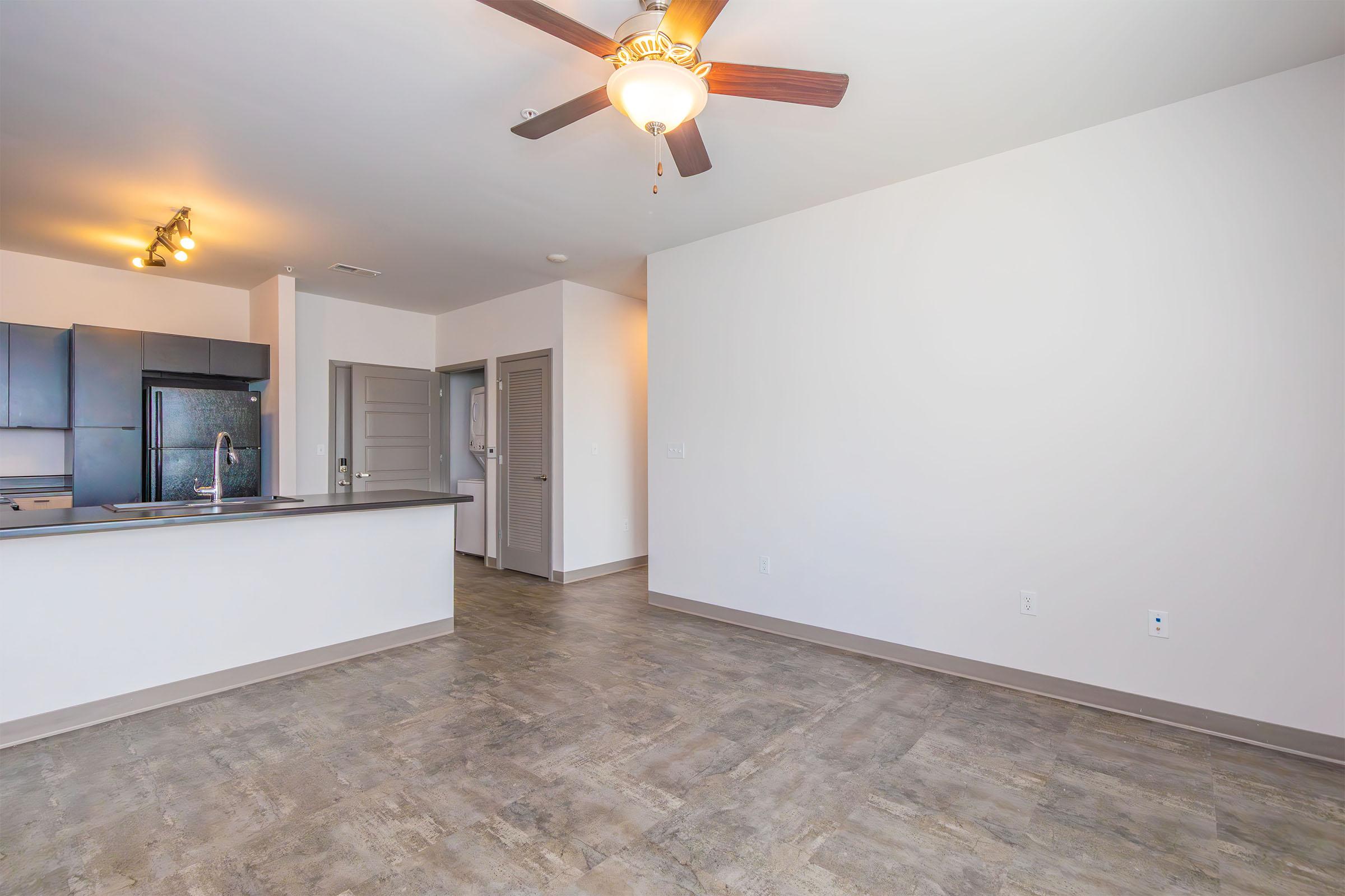 A spacious and modern living area with a ceiling fan, hardwood-style flooring, and a neutral color palette. The open kitchen features dark appliances and a breakfast bar, while a hallway leads to a door on the right, suggesting access to additional rooms. Natural light enhances the inviting atmosphere.