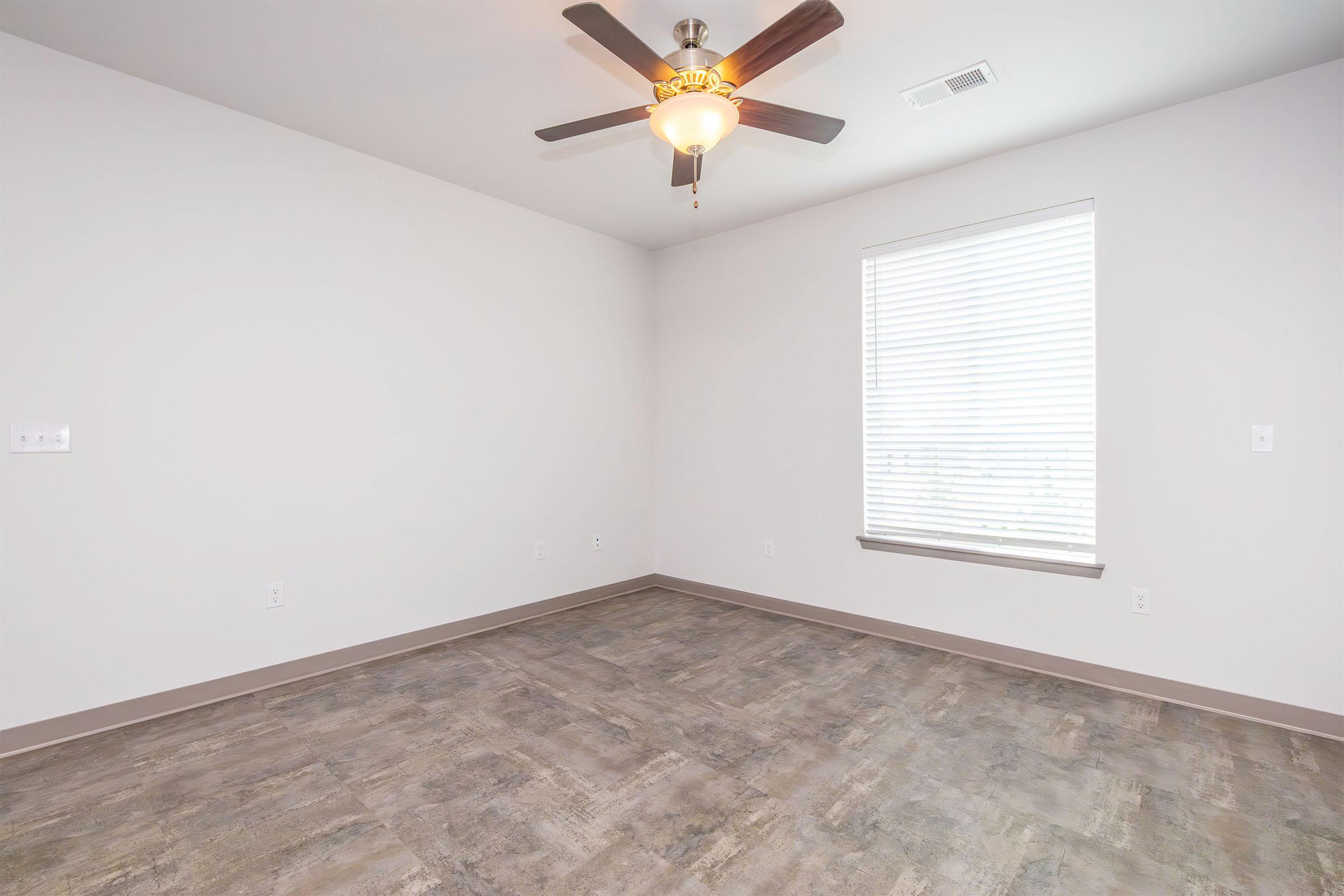 Empty room with light-colored walls, a ceiling fan with a light fixture, and a single window featuring blinds. The flooring is a modern, neutral shade. The space is bright and clean, creating a minimalist atmosphere.