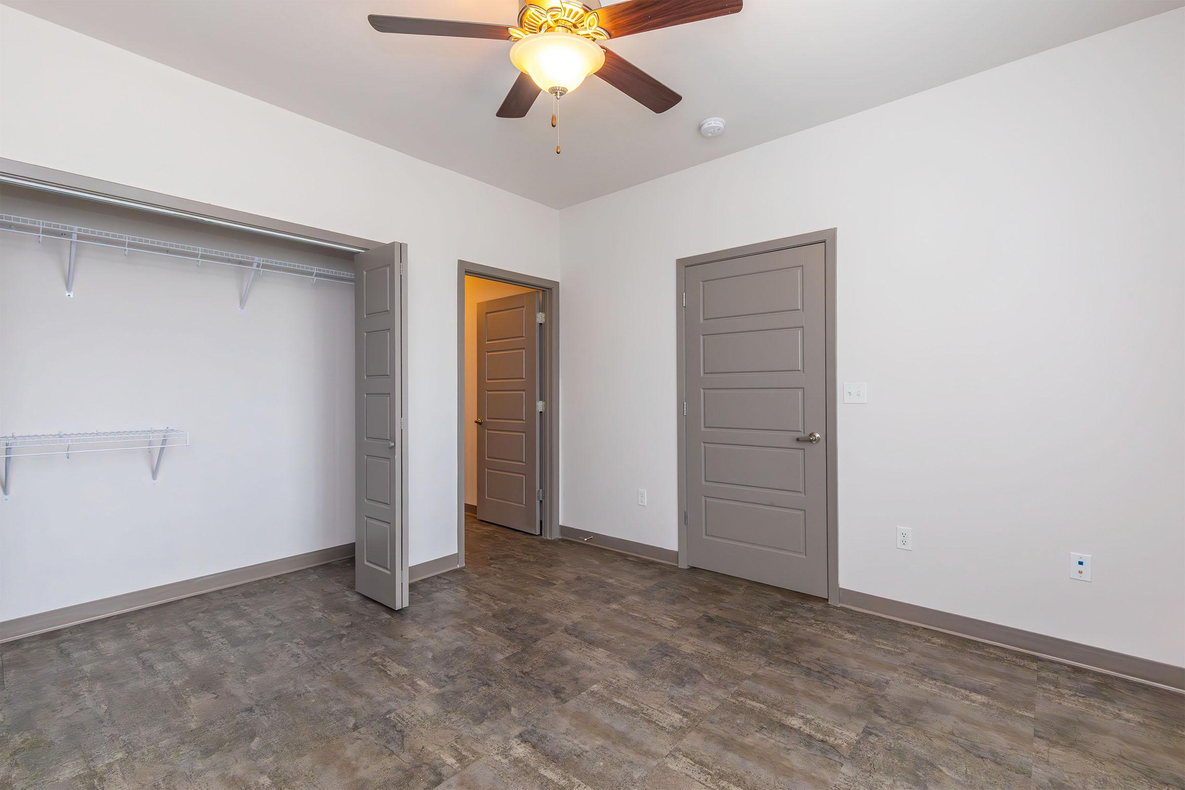 A spacious room featuring a ceiling fan, light-colored walls, and a large closet with shelving. It has two doors leading to other areas and a modern laminate floor. The overall ambiance is bright and welcoming, suitable for various uses such as a bedroom or office space.