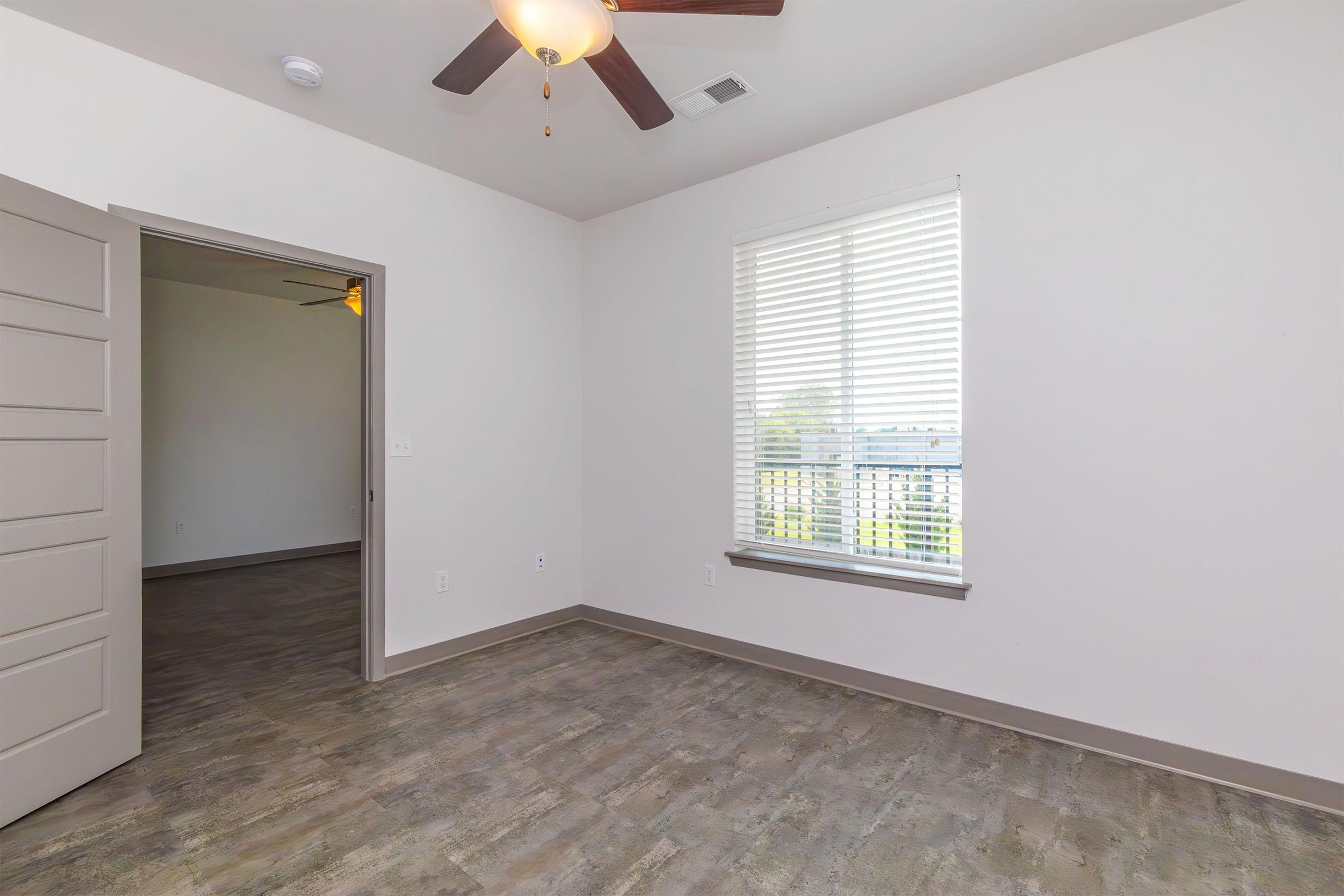 An interior view of a room featuring a ceiling fan, a window with blinds, and a door leading to another space. The floor is made of light-colored wood, and the walls are painted in a neutral shade. The room is well-lit with natural light coming from the window, creating a spacious feel.